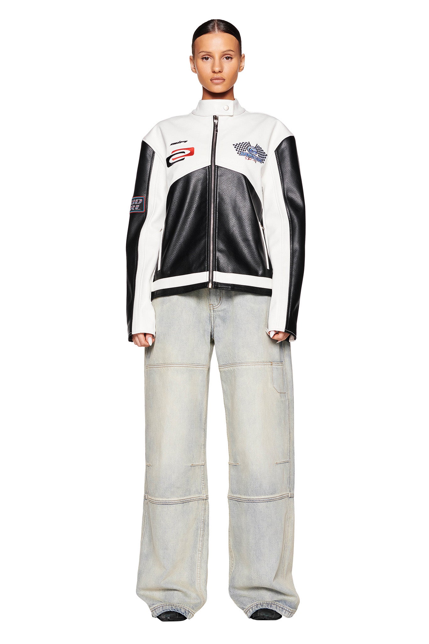 A woman with slicked-back hair is wearing a black and white racing-style I.AM.GIA jacket adorned with patches. She pairs it with the I.AM.GIA MISTY JEAN in blue vintage wash, which are light-colored, wide-leg jeans featuring visible seams. She is standing in a neutral pose against a plain white background.
