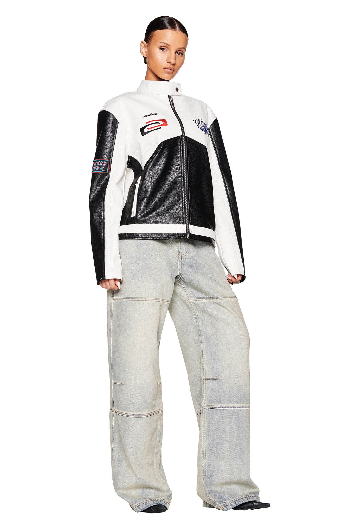 A person stands facing forward, wearing a black and white zip-up jacket with red and black patch designs from I.AM.GIA. They are also sporting the MISTY JEAN - BLUE : VINTAGE WASH from the same brand, along with white sneakers. Their hair is neatly pulled back against the plain white background.