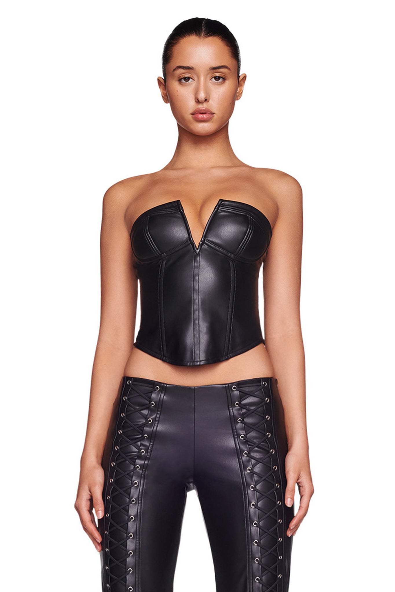 A woman stands against a white background wearing the EVANNA CORSET - BLACK from I.AM.GIA, a strapless leather top with a plunging neckline, paired with matching high-waisted leather pants featuring lace-up detailing along the front sides. Her hair is pulled back, and she faces forward, capturing the edgy aesthetic of I.AM.GIA.