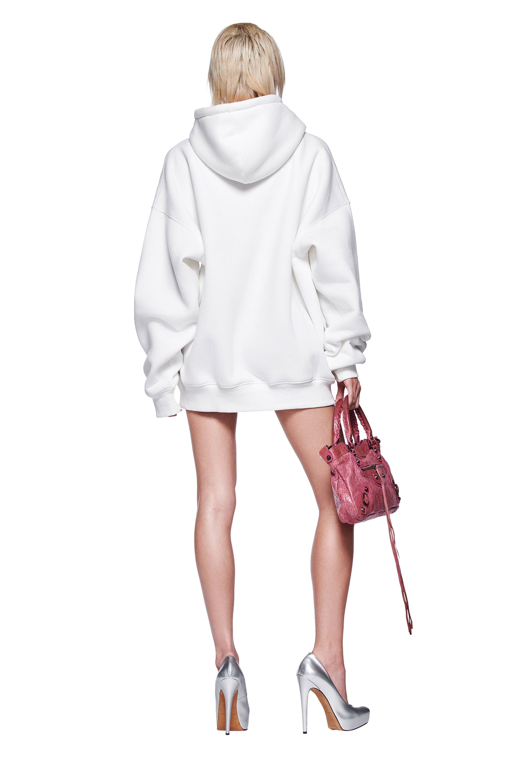 A person with short blonde hair faces away, wearing the I.AM.GIA DORIAN HOODIE in white, styled with silver heels and a small pink handbag against a plain white background.