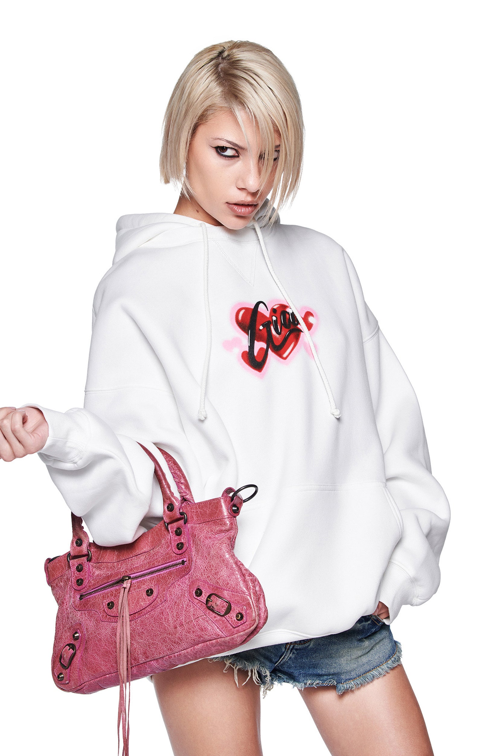 A person with short blonde hair wears the oversized I.AM.GIA DORIAN HOODIE in white, featuring a heart and "Love" airbrush art, styled with denim shorts and a pink handbag against a white background.