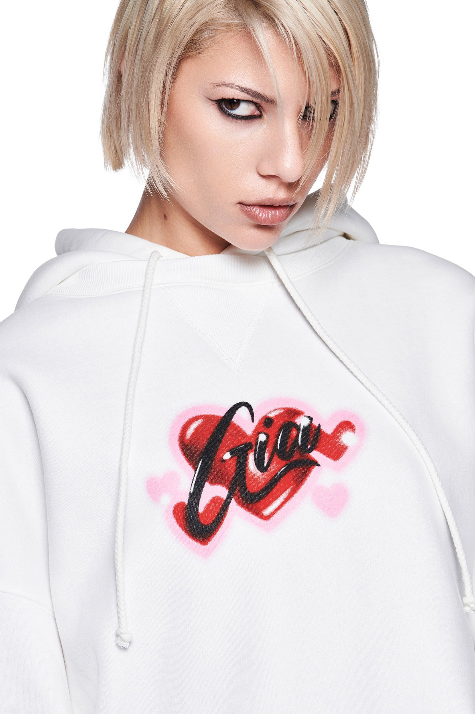 A person with short blonde hair and bold eye makeup wears the oversized I.AM.GIA DORIAN HOODIE in white, featuring a red and pink heart graphic and "Gia" in black airbrush script across the chest.