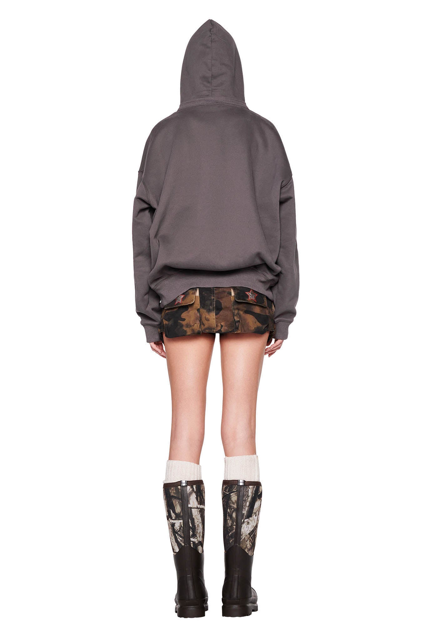 A person stands facing away from the camera, wearing the DORIAN HOODIE - GREY : CHARCOAL by I.AM.GIA with the hood up, camo print shorts, tall camo print boots, and white knee-high socks. The background is white.