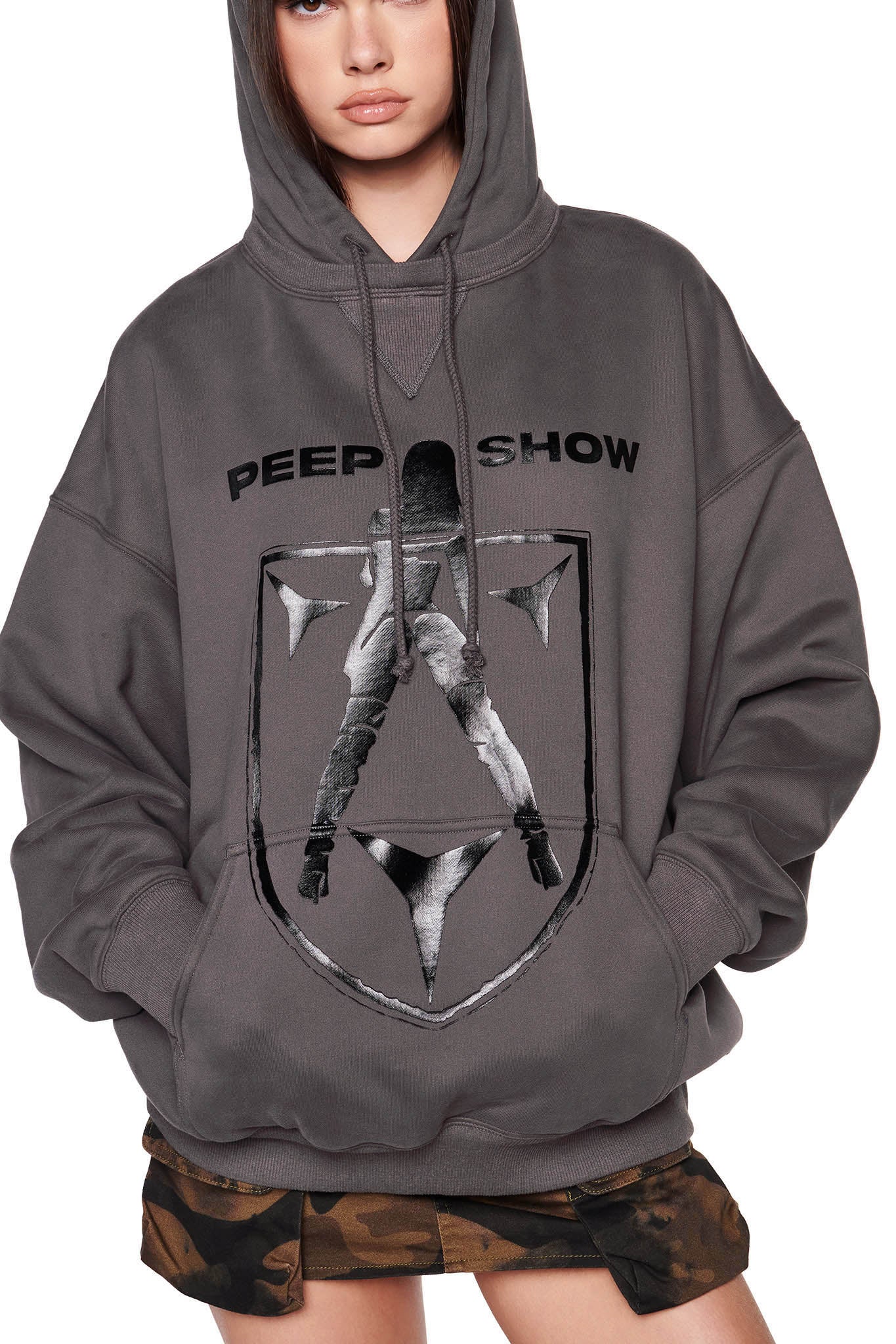 A person wearing an oversized charcoal DORIAN HOODIE - GREY from I.AM.GIA stands against a white background. The hoodie features the text "PEEP SHOW" and a graphic of a dancer with their back turned. The person is also sporting camo-patterned shorts.