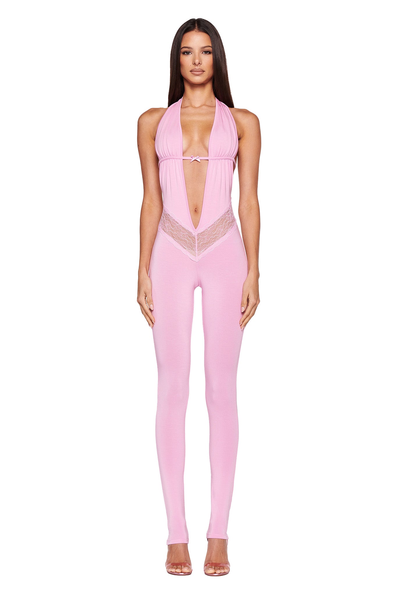 A person with long hair is wearing the pink, form-fitting ASTORA JUMPSUIT from I.AM.GIA. The sleeveless outfit features lace details and cutouts on the chest and waist. They are standing with their arms at their sides against a plain white background and are paired with high-heeled, clear shoes.