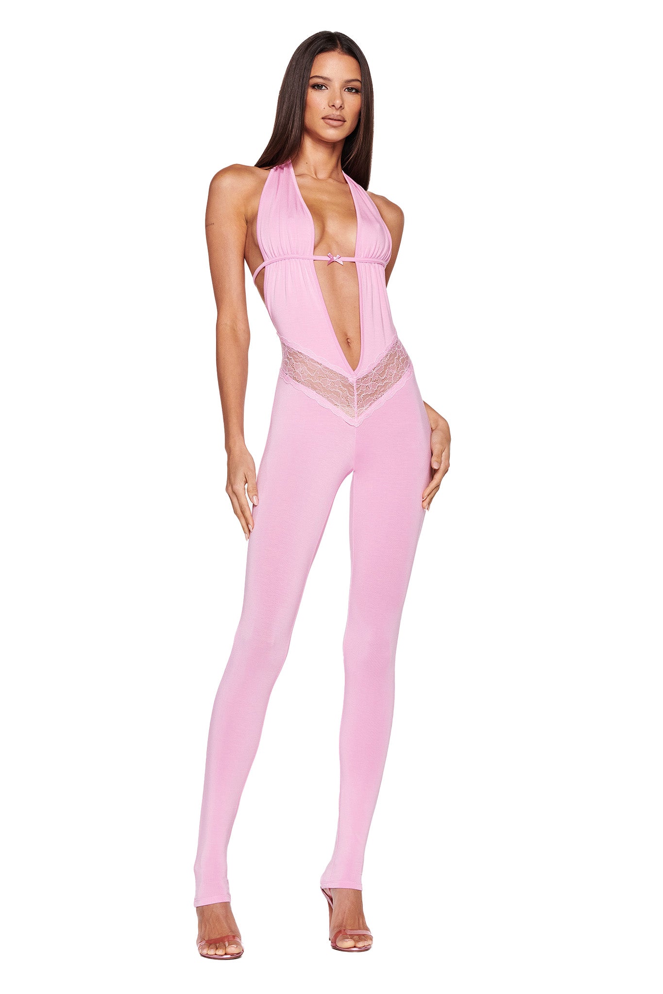 A person with long hair is posing in an I.AM.GIA ASTORA JUMPSUIT - PINK, featuring a halter-neck design with lace details on the sides and a plunging neckline. They are wearing clear high-heeled shoes against a plain white background.