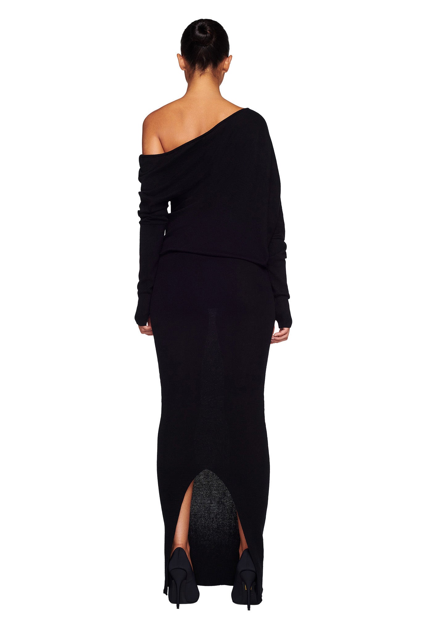 A person with dark hair is standing with their back to the camera, wearing a chic black GISELLE MAXI DRESS by I.AM.GIA. The sleek design features an off-the-shoulder neckline and long sleeves, complemented by a slit at the back. The ensemble is completed with striking black high heels.