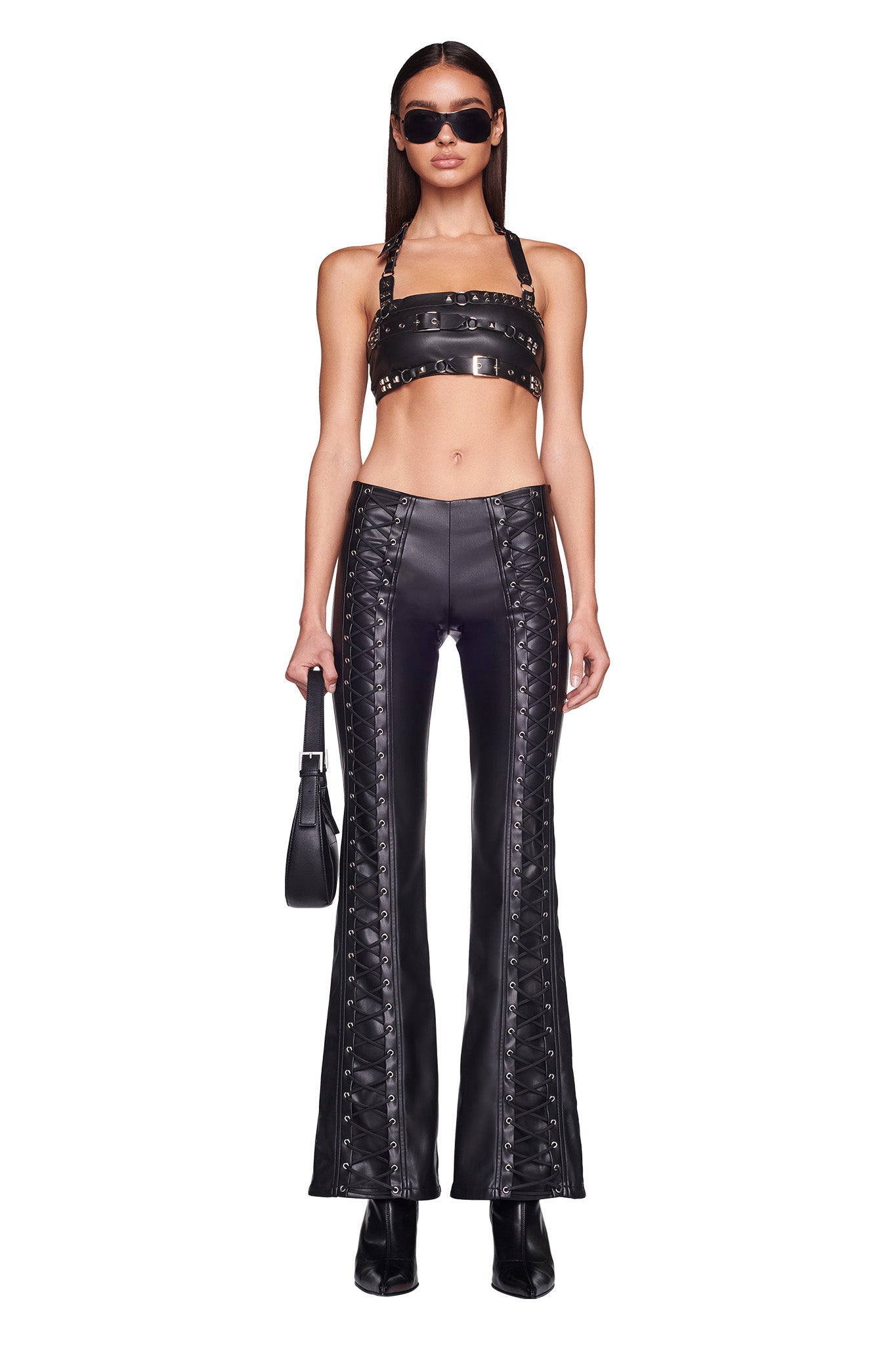 A woman stands confidently in an I.AM.GIA black leather crop top with metal details, paired with the I.AM.GIA SONORA PANT - BLACK, which are high-waisted flared leather pants adorned with laces on the sides. She accessorizes with black sunglasses, a black handbag, and black boots, posing against a white background.