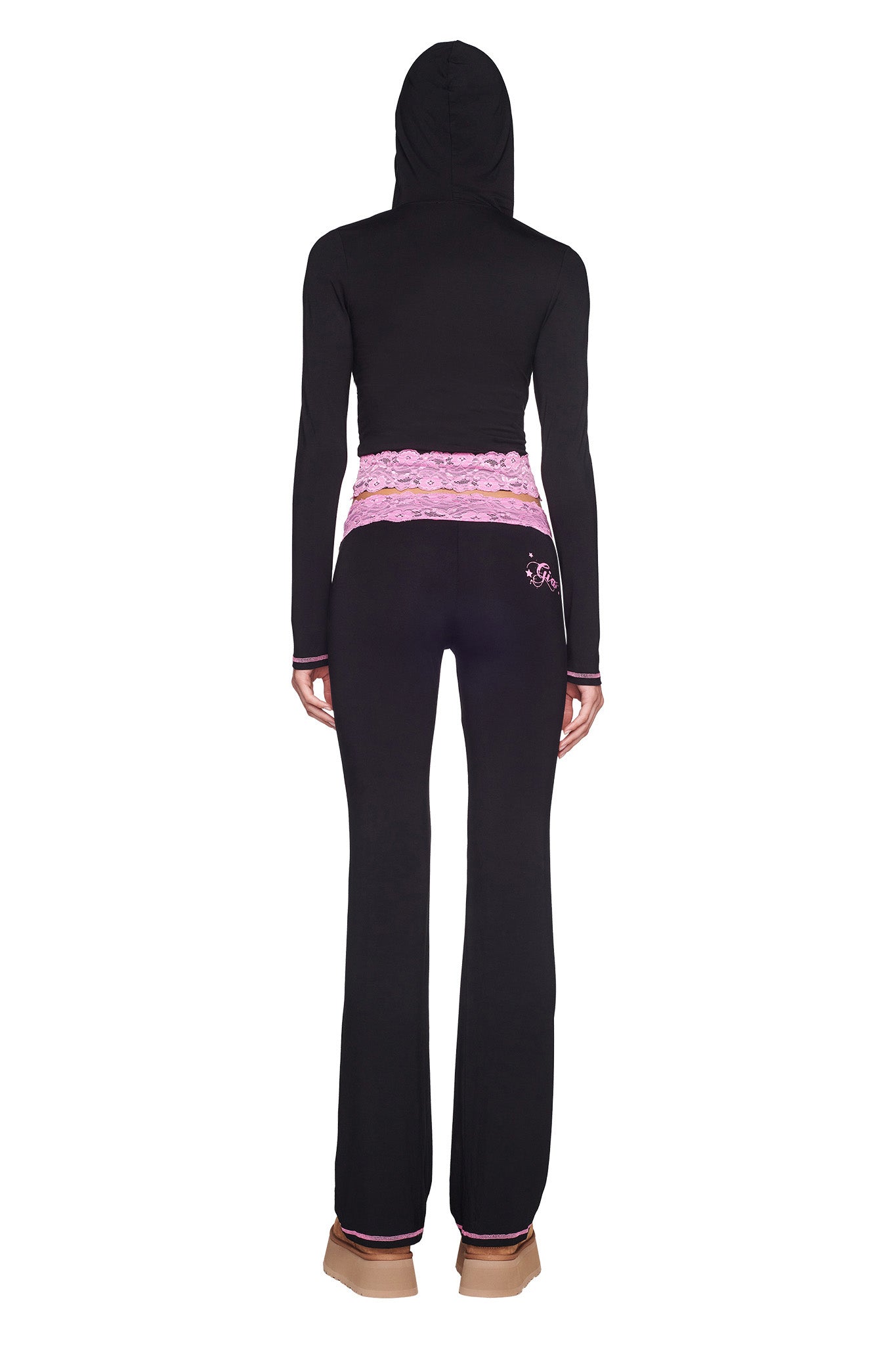 A person stands facing away from the camera, donning an I.AM.GIA Tarsha Jacket in Black/Pink along with matching pants that have a pink lace waistband. The outfit features subtle pink accents, and the individual is also wearing light brown shoes.