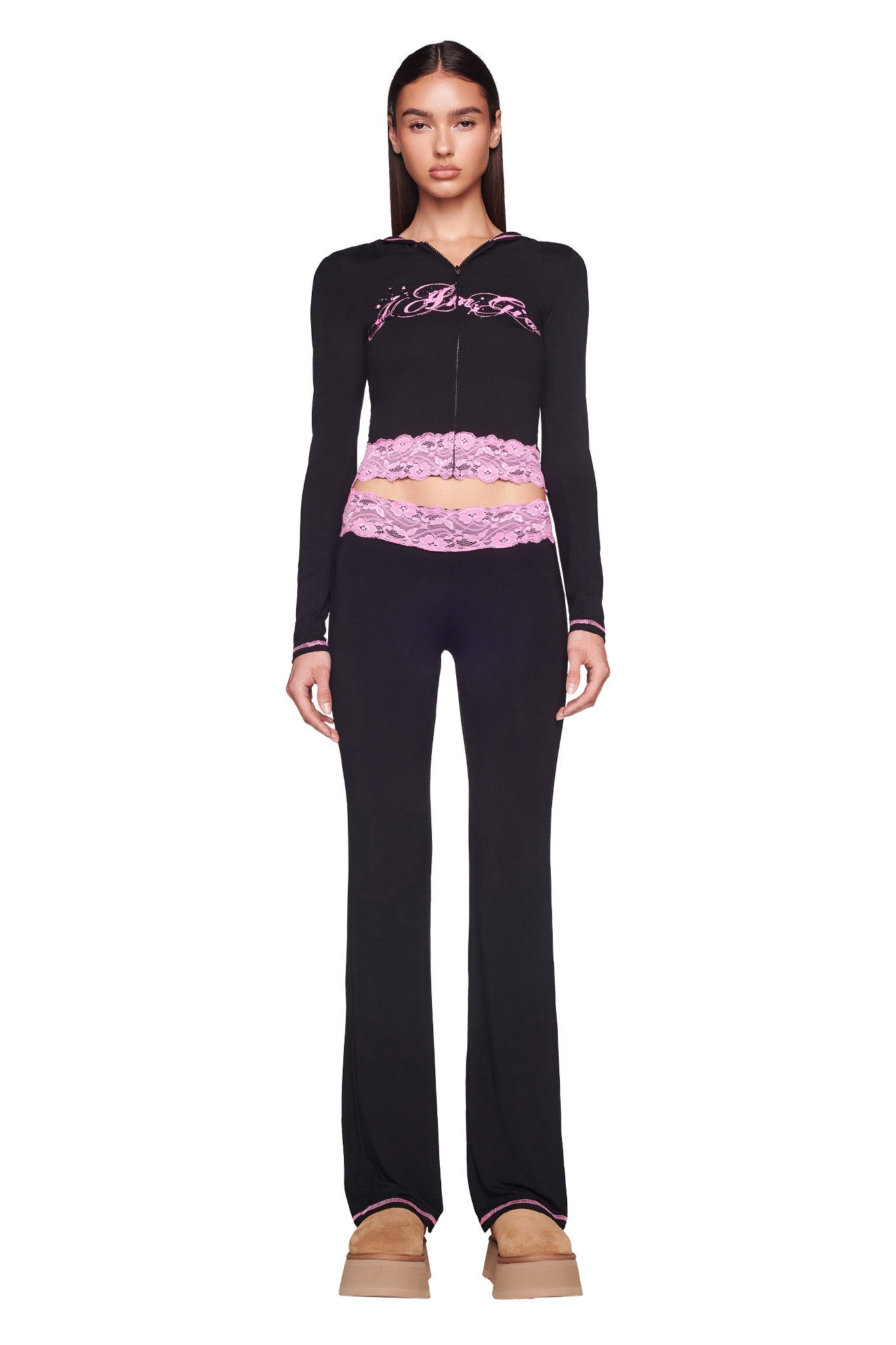 A person with long dark hair is wearing a black and pink I.AM.GIA Tarsha Jacket, black pants with pink lace at the waistband, and tan platform shoes. They are standing against a plain white background.