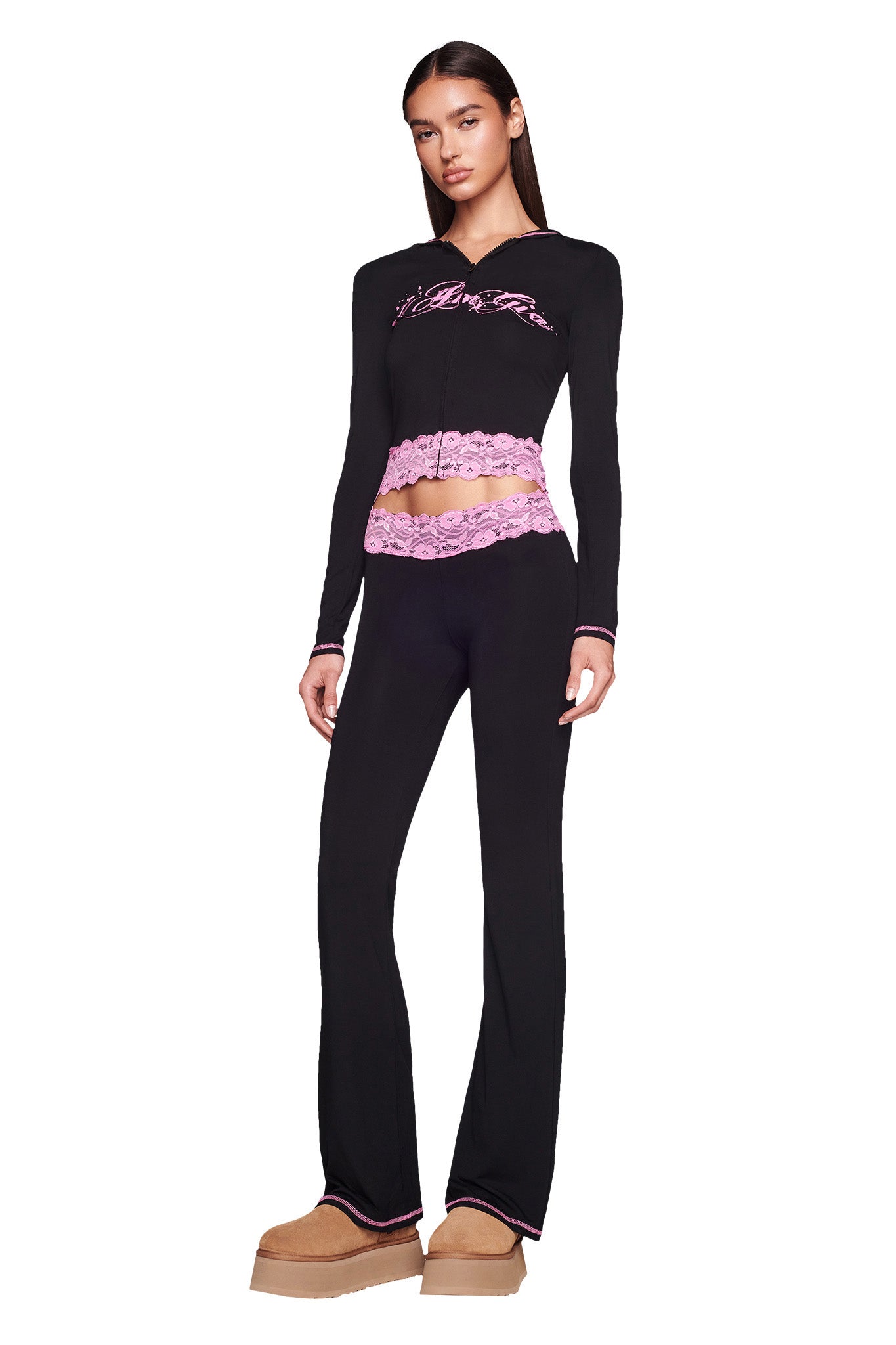 A woman stands confidently wearing the TARSHA JACKET in black with pink lace trim from I.AM.GIA, paired with matching black pants. She is also wearing beige platform shoes. Her long dark hair is straight and loose, and her facial expression is neutral against the white background.