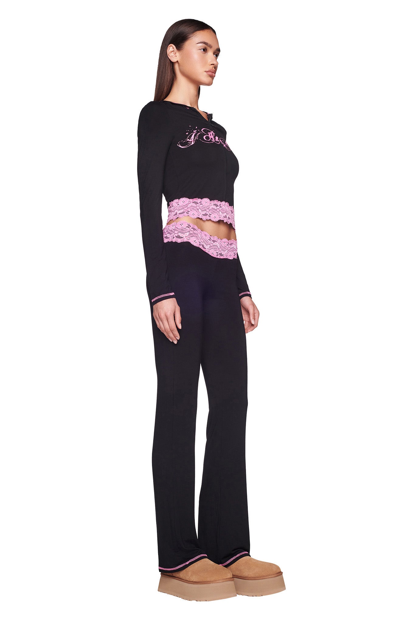 A woman with long hair stands in profile, wearing the TARSHA JACKET in black and pink from I.AM.GIA, paired with matching black pants. She is also wearing tan platform shoes. The jacket features decorative elements around the midsection and chest. The background is plain white.
