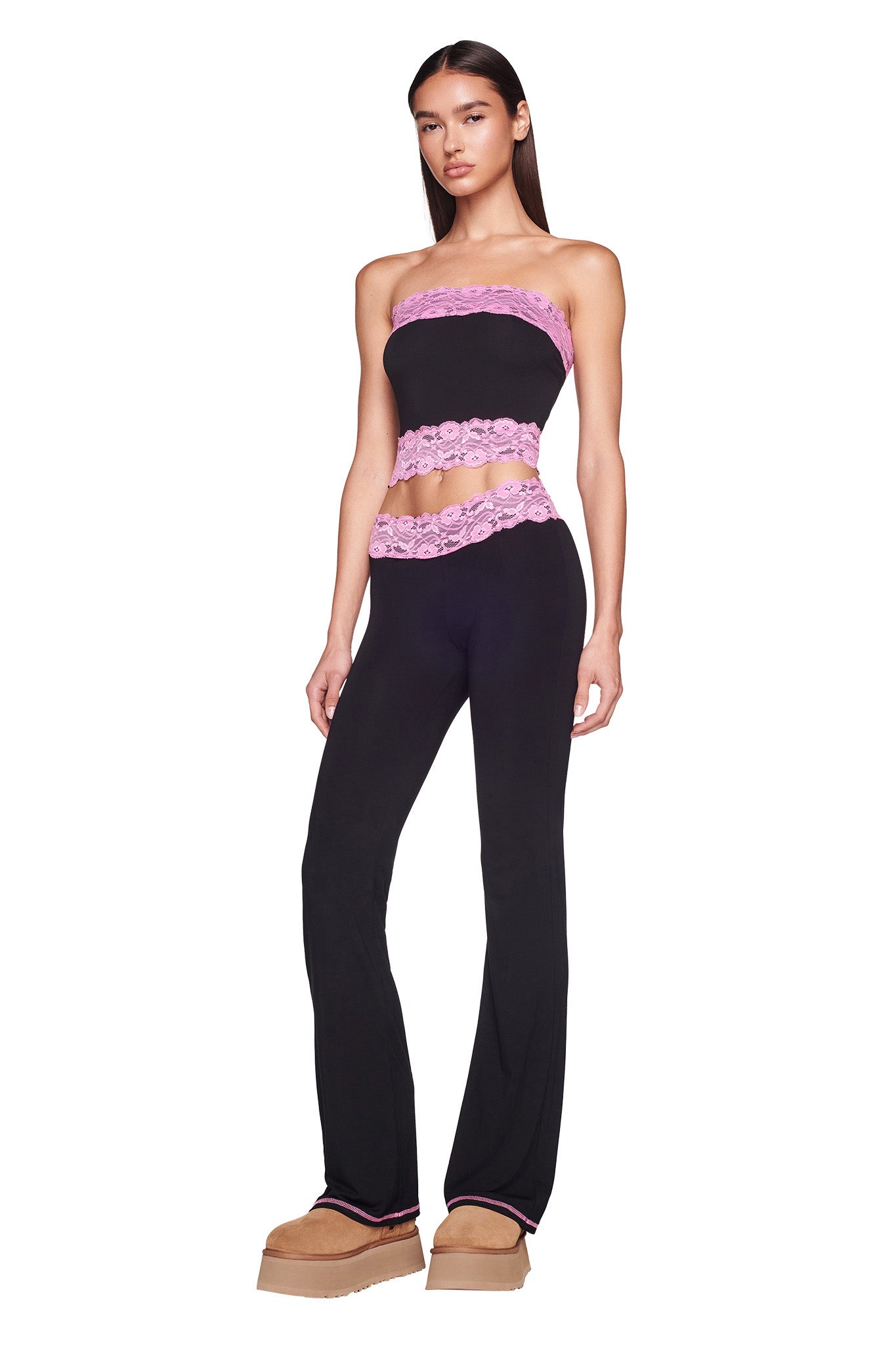 A woman stands wearing the TARSHA PANT in black with pink lace trim from I.AM.GIA, paired with a matching strapless black tube top also adorned with pink lace trim. She complements her outfit with tan platform shoes. Her long, straight hair cascades down her back as she poses against a white background.