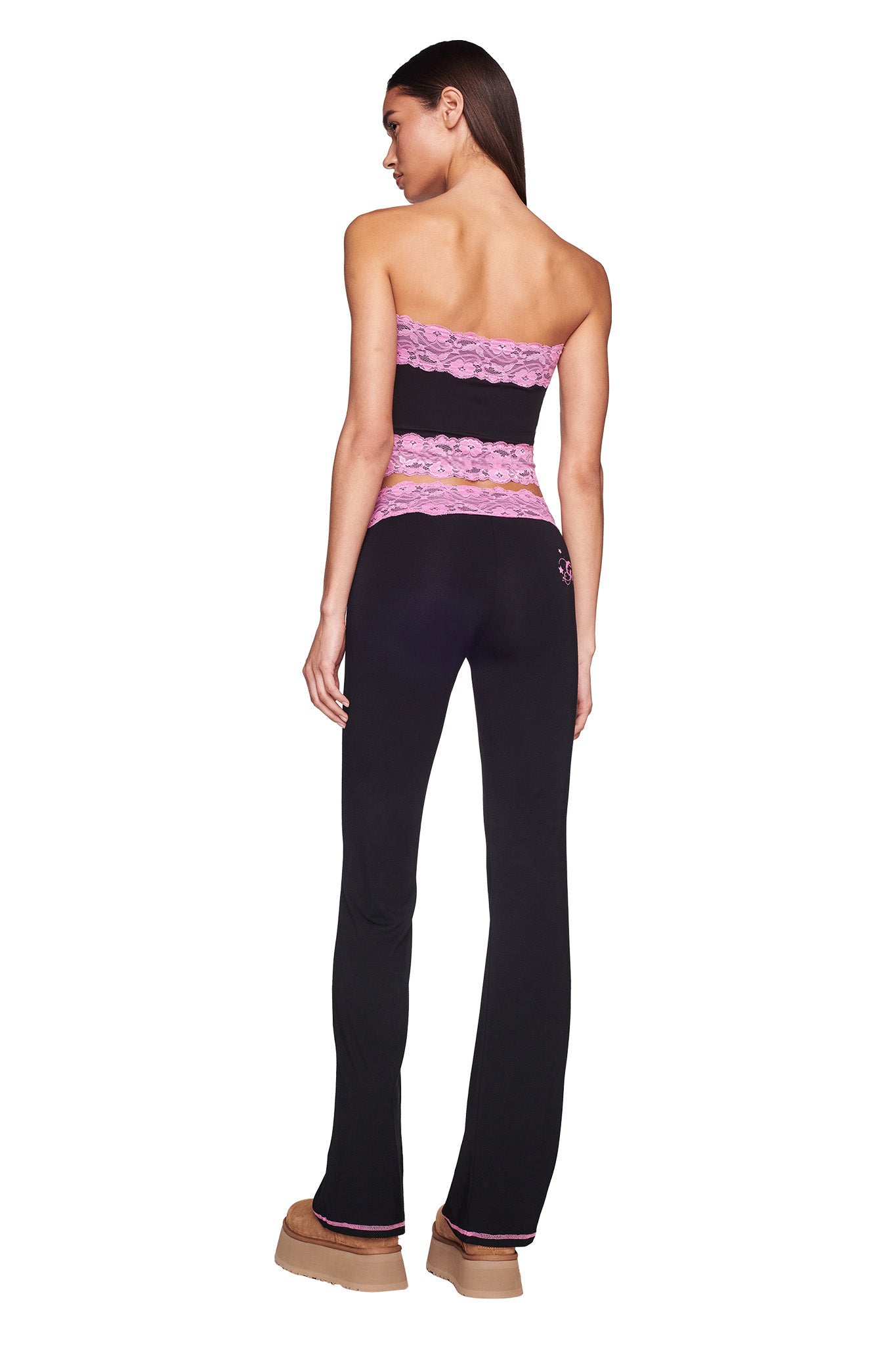 A woman stands facing away, wearing the TARSHA BOOB TUBE - BLACK/PINK from I.AM.GIA, a strapless top with floral lace details. She pairs it with matching black pants featuring a pink logo near the hip and tan platform shoes. Her long, dark hair flows down her back.