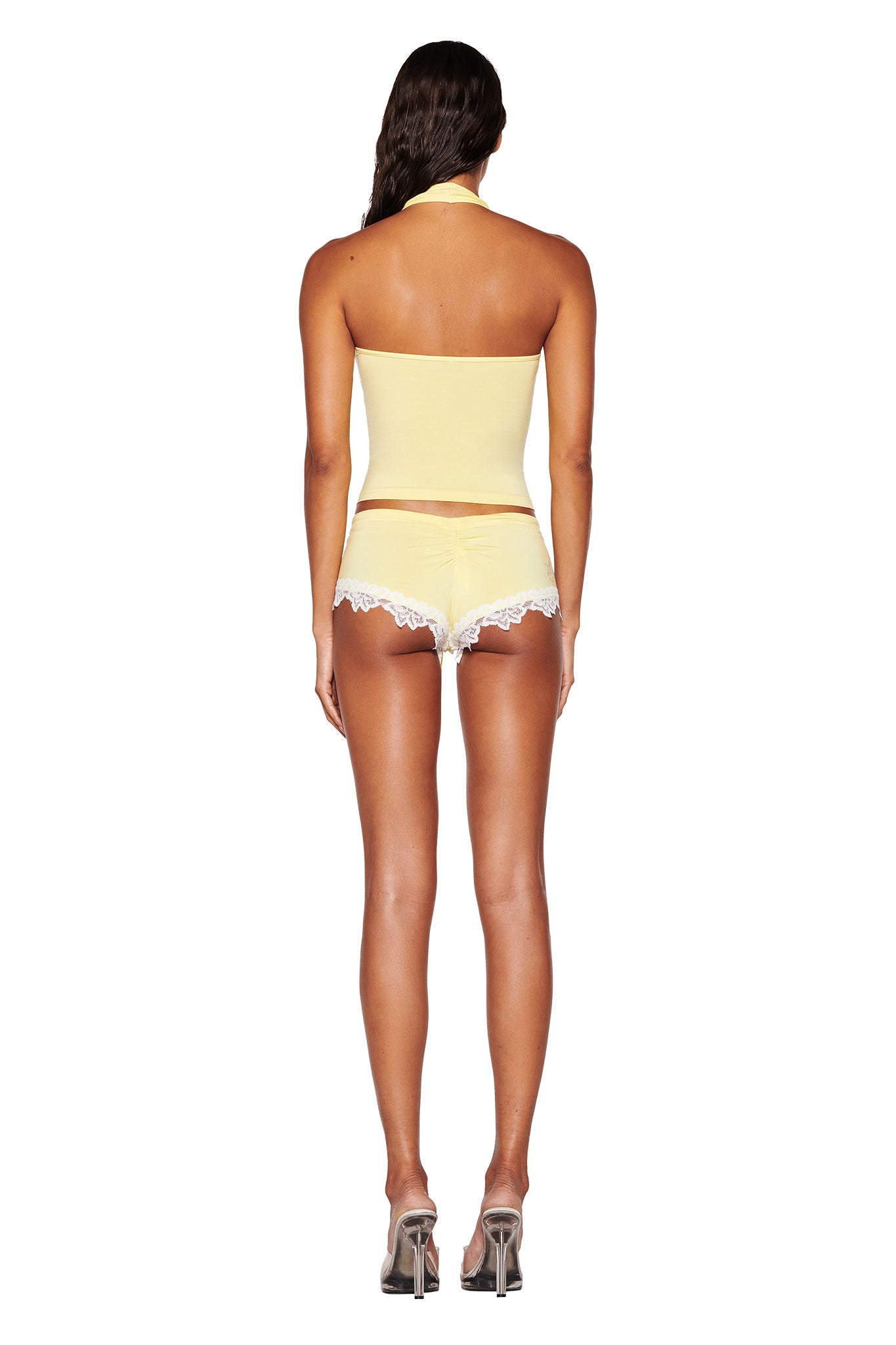 Standing with their back turned, a person with long hair wears a baby yellow tube top perfectly paired with the CELESTE SHORT - YELLOW: BABY YELLOW by I.AM.GIA, accented by white lace trim. The outfit, boasting a subtle bodycon fit, is enhanced by high-heeled shoes against a plain white backdrop.