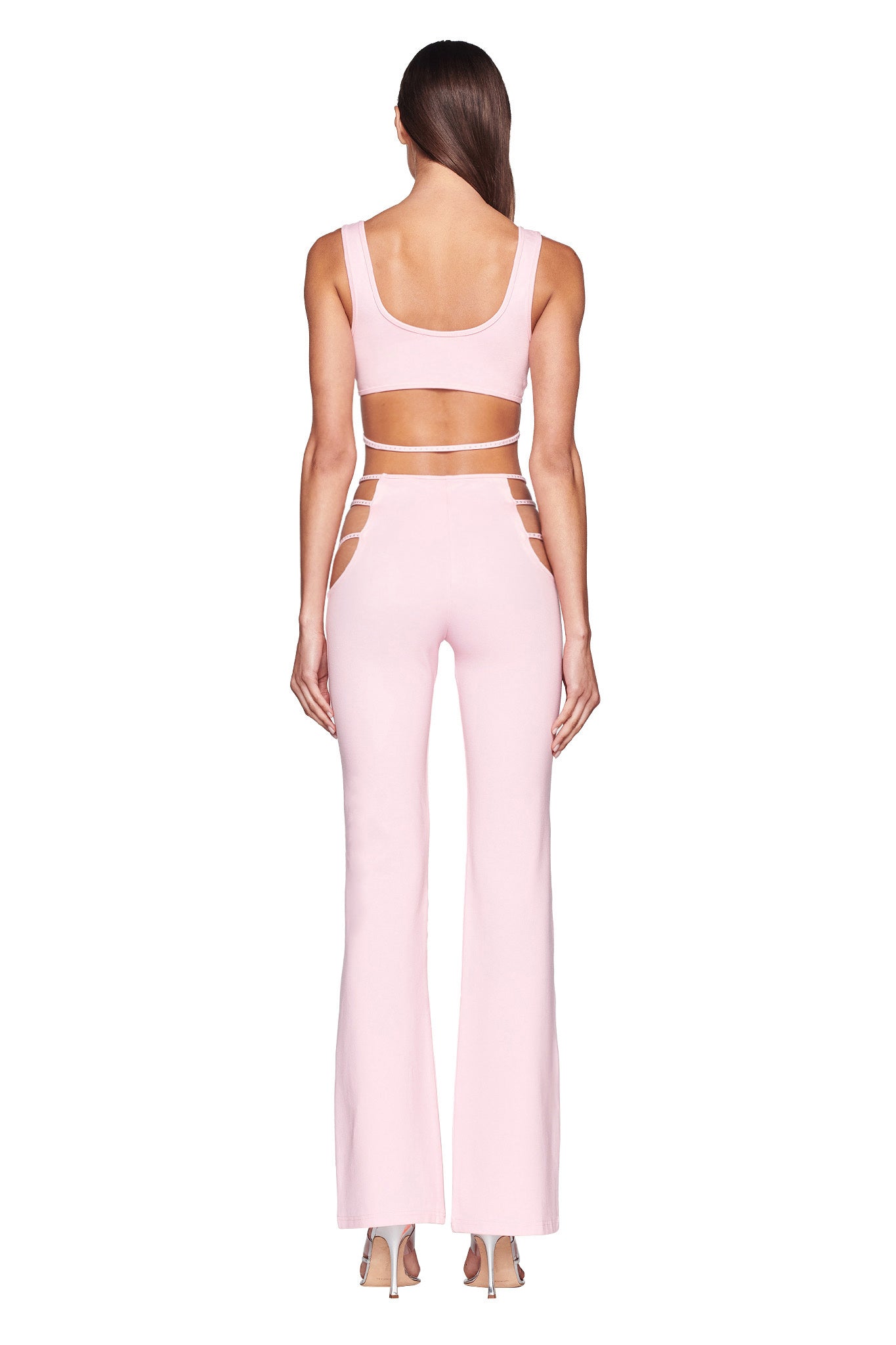 A person stands facing away from the camera wearing an I.AM.GIA light pink outfit. The sleeveless top has a low scoop back, and the high-waisted ROXANNA PANTS - PINK : BABY PINK feature cutouts on the sides near the hips. The person is wearing high-heeled shoes, and their long hair is loose down their back.