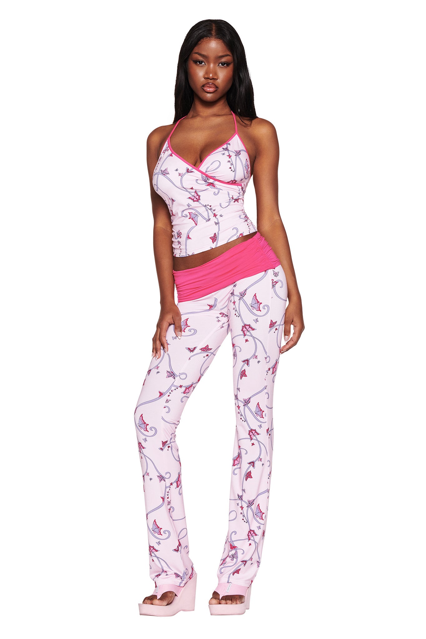 A person models the I.AM.GIA JANNIS PANT - PINK : BUTTERFLY RIBBON ensemble on a white backdrop. The sleeveless top and matching pants feature a pink and white floral pattern with butterfly ribbon accents, paired with platform sandals for a bold look.