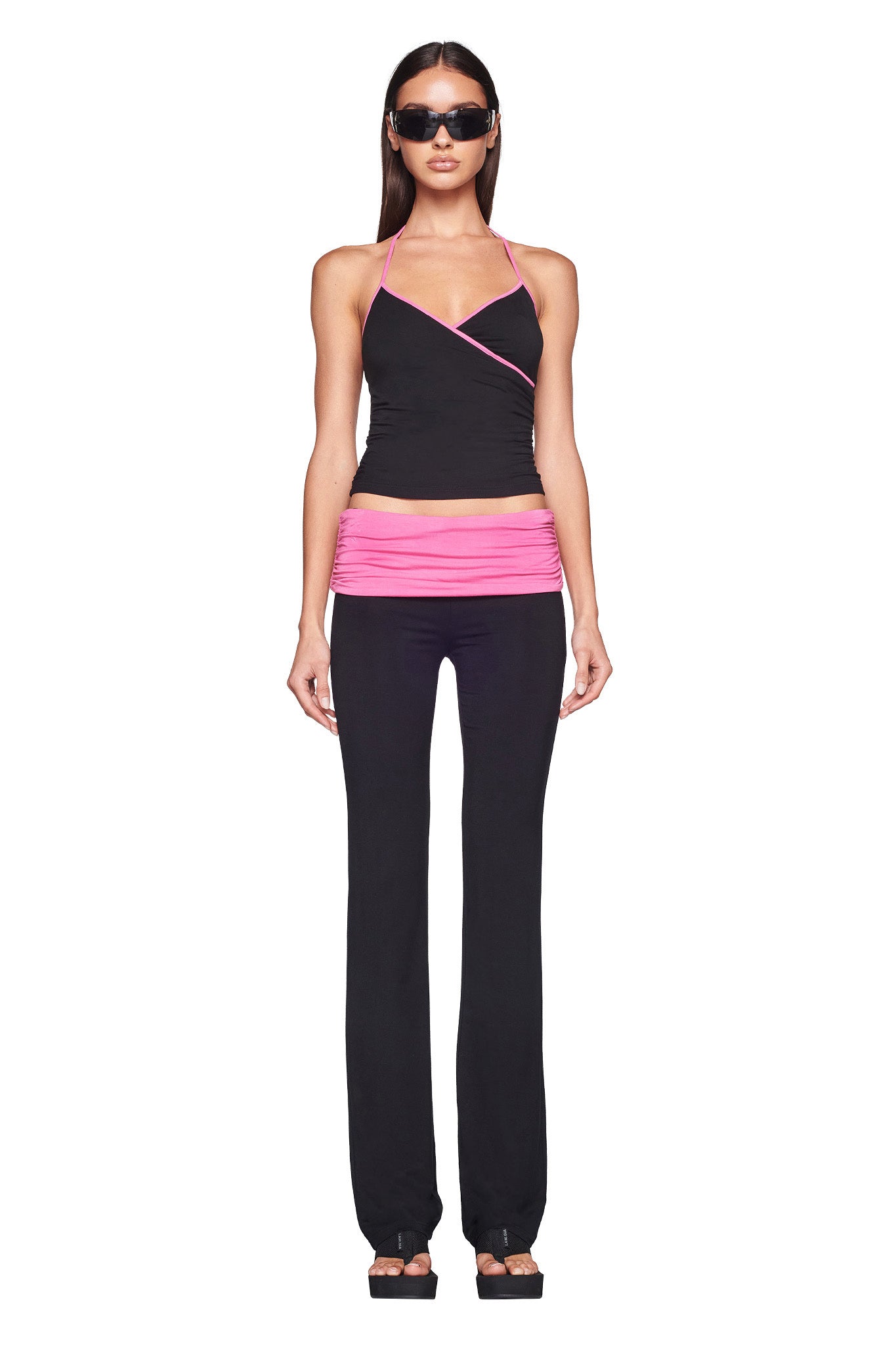 A woman is standing against a plain white background, wearing the I.AM.GIA JANNIS PANT in black and pink paired with a matching top featuring halter neck and wrap details. She has long hair and is accessorized with dark sunglasses and black heeled sandals.