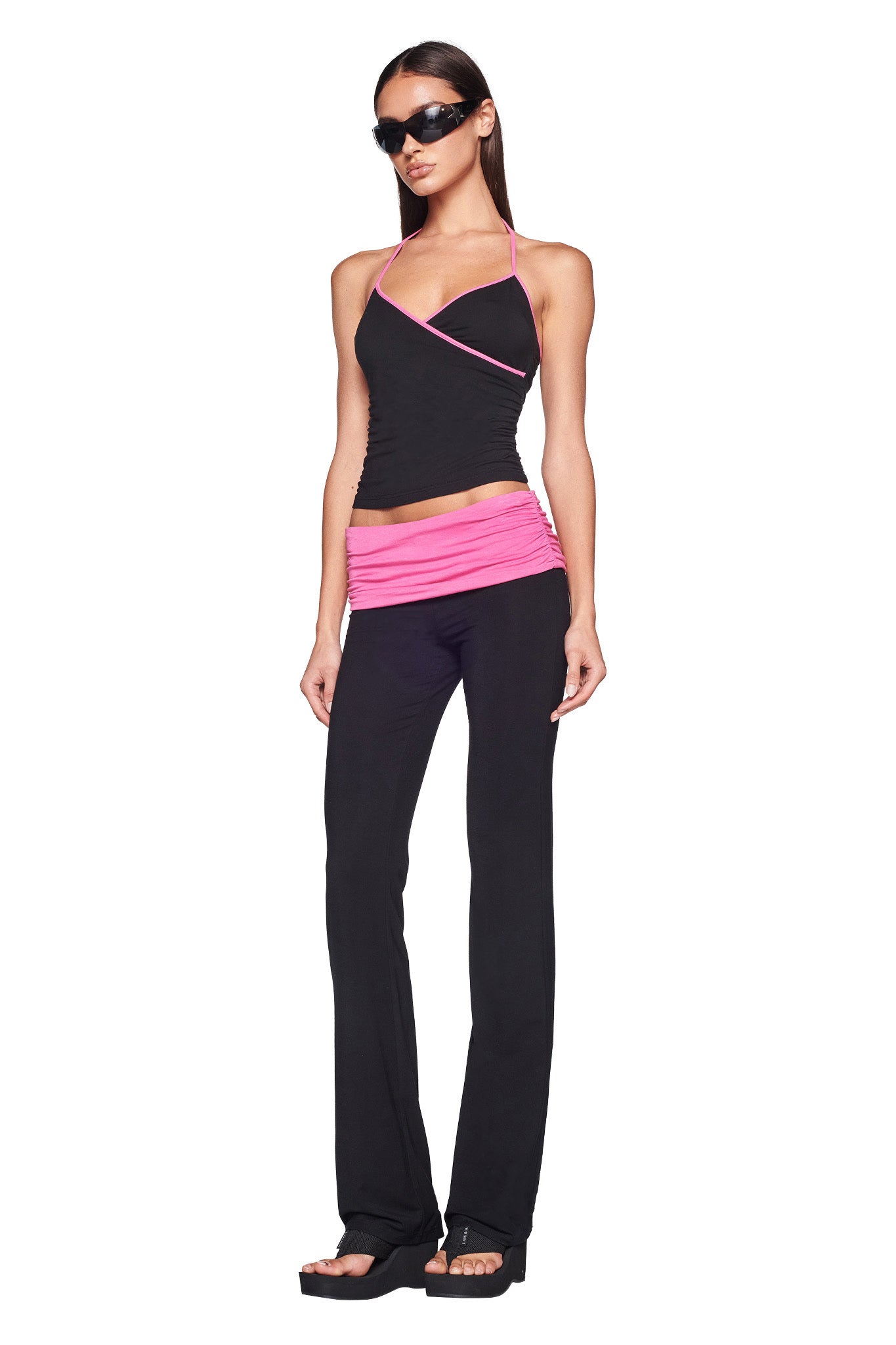 A woman with long, dark hair is wearing dark sunglasses, a black I.AM.GIA halter top with pink accents, the JANNIS PANT - BLACK/PINK by I.AM.GIA featuring a pink waistband, and black platform sandals. She stands confidently against a white background.
