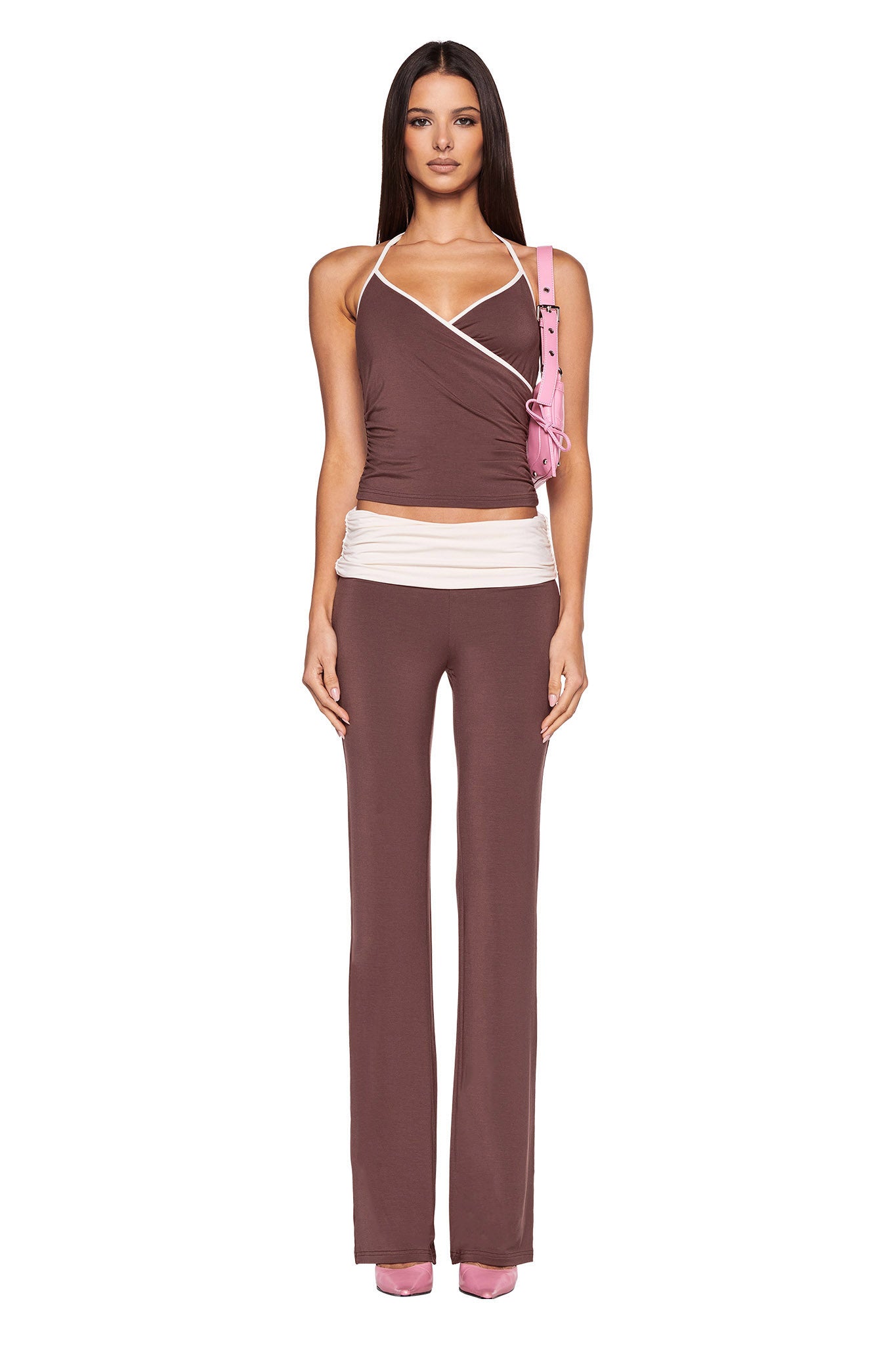 A woman stands facing forward, dressed in a brown halter top with white accents paired with the I.AM.GIA JANNIS PANT - BROWN : CHOCOLATE, featuring a white waistband. She complements her outfit with pink shoes and a matching pink shoulder bag. Her long, dark hair is straight and loose against the plain white background.