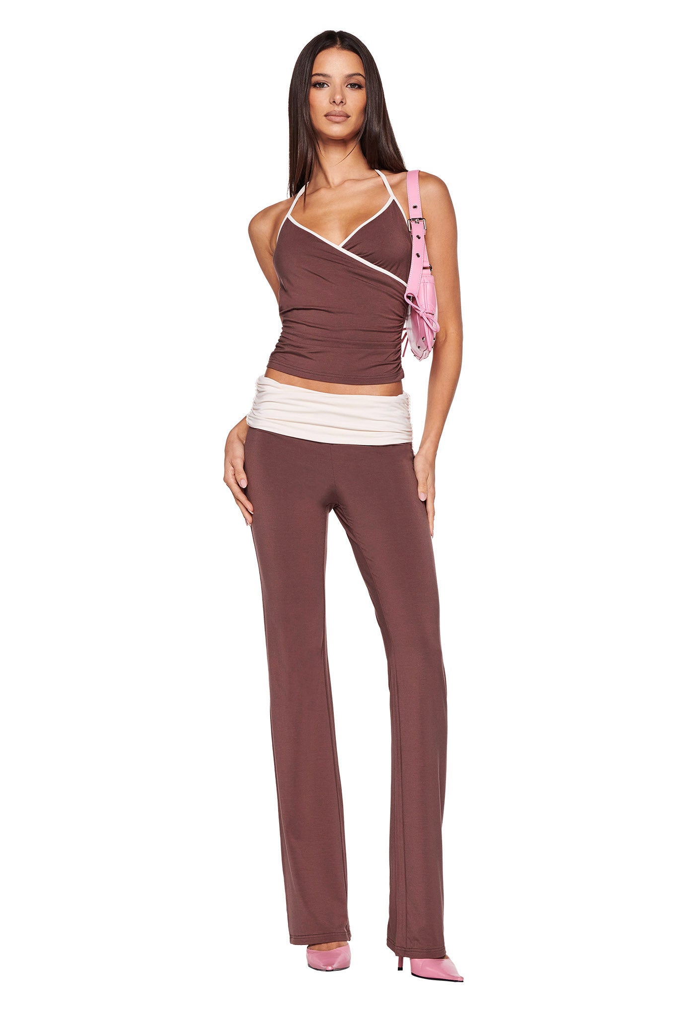 A woman stands confidently, wearing a stylish brown halter top with white trim, paired with I.AM.GIA’s JANNIS PANT in a rich chocolate colorway. She accessorizes with pink high heels and a small pink shoulder bag while posing against a plain white background.