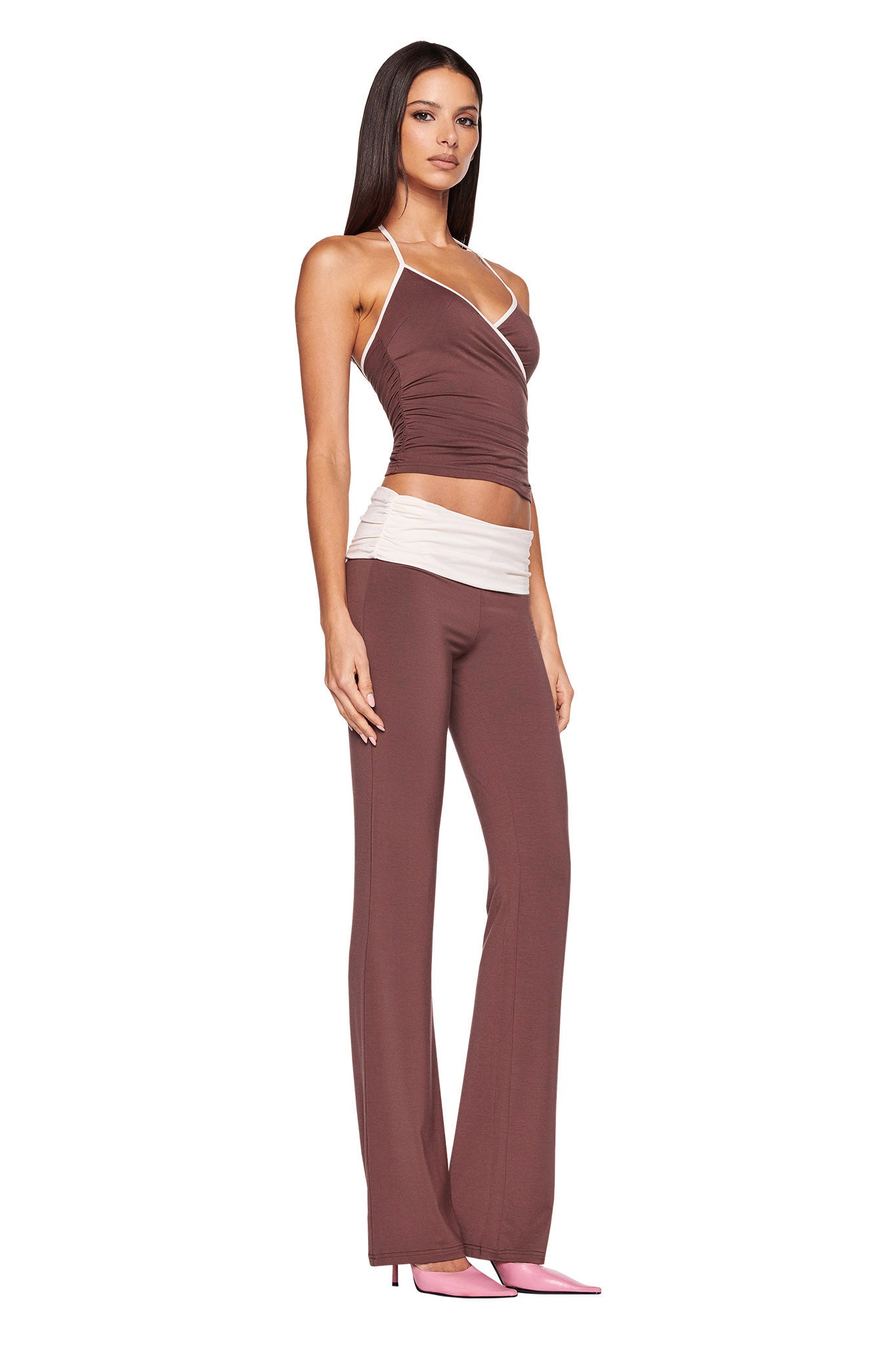 A woman with long brown hair is wearing a fitted, sleeveless top in a rich brown chocolate colourway, paired with the JANNIS PANT - BROWN : CHOCOLATE from I.AM.GIA and pink high-heeled shoes. She stands with one arm by her side and the other behind her back, gazing toward the camera.