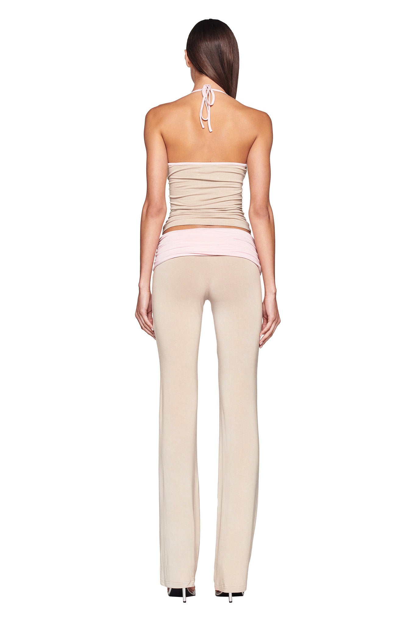 A person with long, straight hair is shown from the back, wearing an I.AM.GIA JANNIS TANK in neutral tan/pink that ties at the neck and matching beige I.AM.GIA flared pants. They are accessorized with a pink waistband around their hips. The background is plain white.