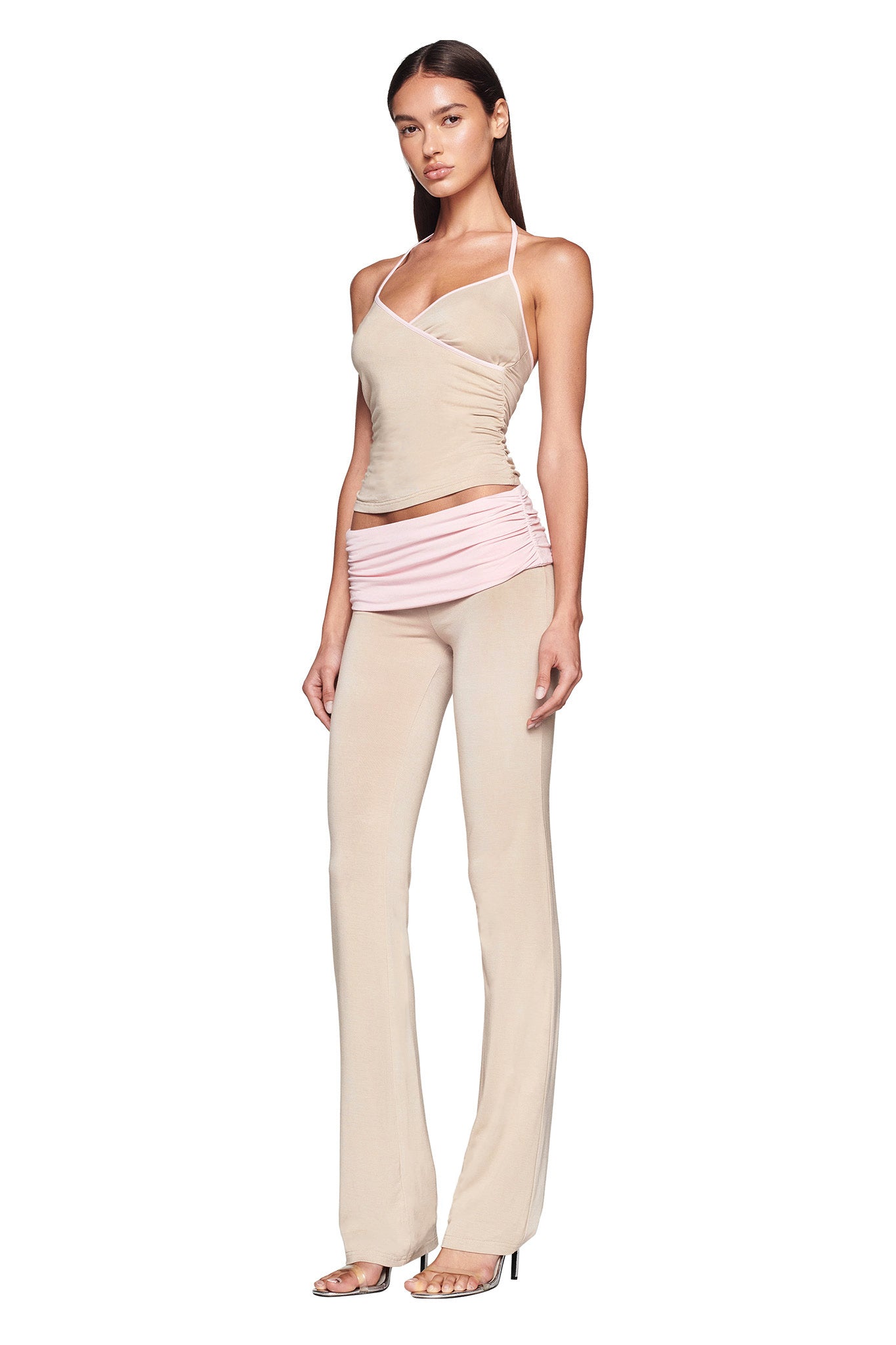 A woman stands against a white background, wearing the JANNIS TANK - NEUTRAL : TAN/PINK from I.AM.GIA, a beige form-fitting sleeveless top with asymmetric straps, paired with high-waisted pants in beige that feature pink ruched detailing around the hips. She completes the look with high-heeled sandals.