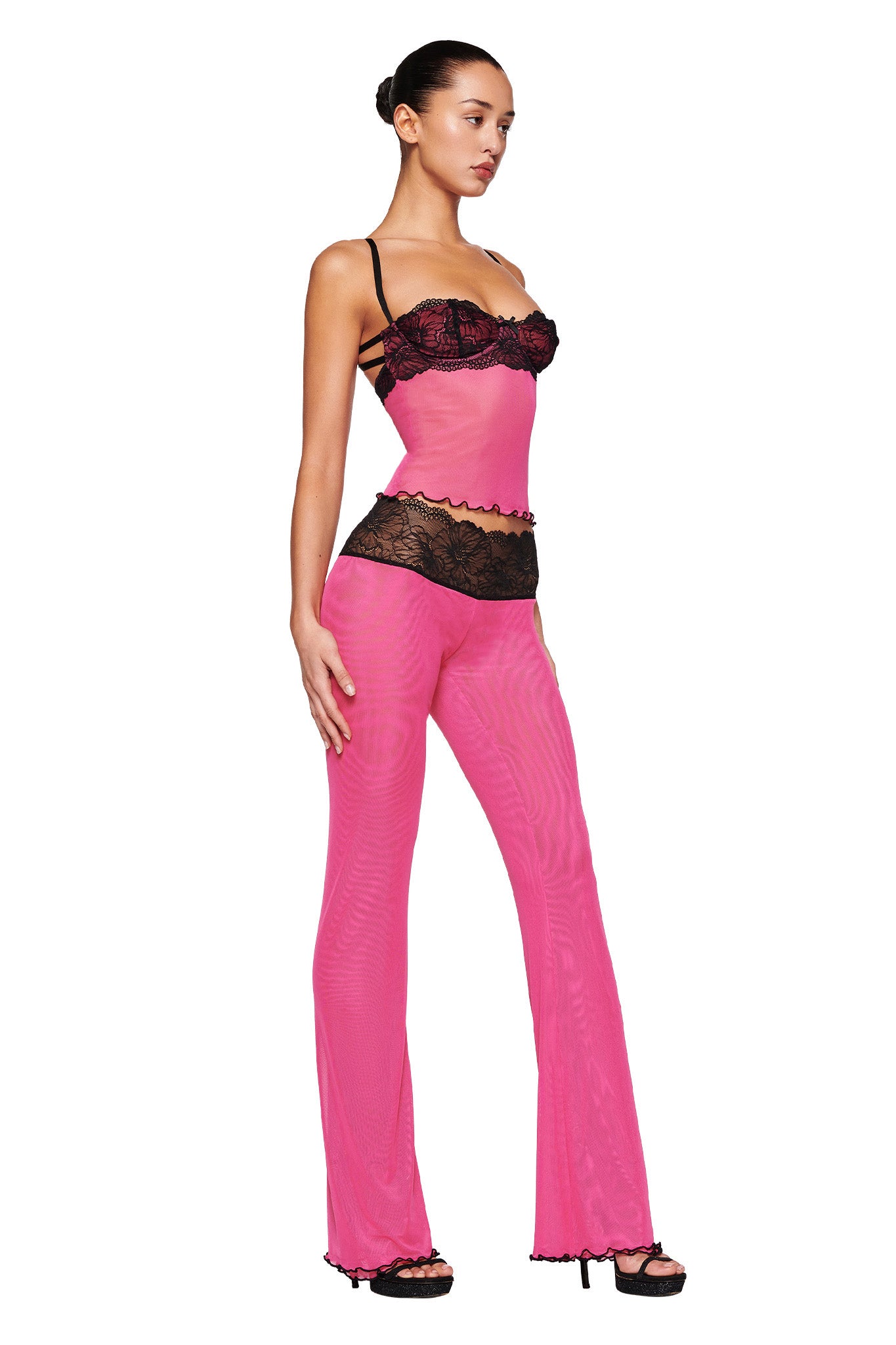 Someone is shown in profile wearing the I.AM.GIA SILO TOP - PINK, a sheer, vibrant pink two-piece outfit with black lace details. The mesh fabrication includes a fitted top with spaghetti straps and matching high-waisted pants. Standing confidently with their hair pulled back and high-heeled sandals, they exude style and poise.