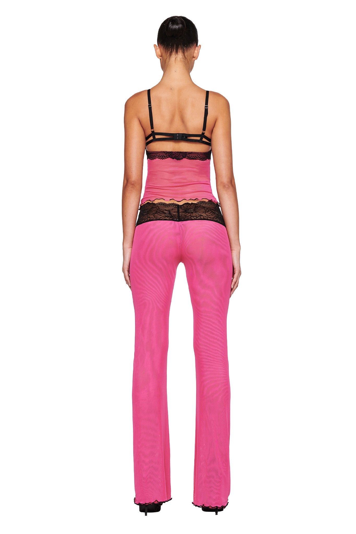A person with long hair tied back is wearing the I.AM.GIA SILO TOP in pink, featuring sheer fabric with black straps and lace details. They have paired it with matching pink sheer pants that have black lace accents on the waistband. The person is facing away from the camera, highlighting the intricate mesh fabrication of their outfit.
