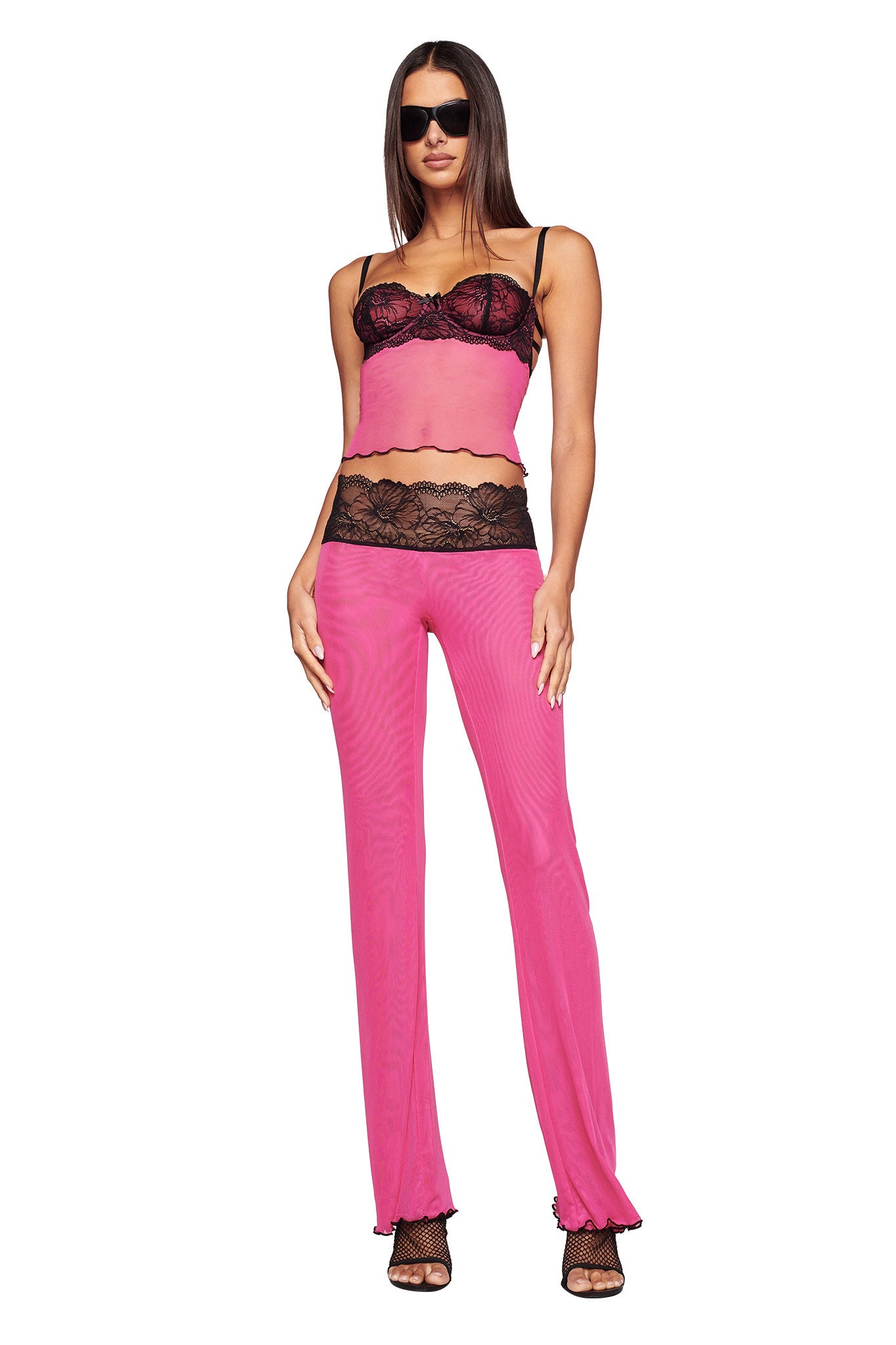 A woman wearing the I.AM.GIA SILO TOP in pink, featuring spaghetti straps and black lace accents at the waist and bust, with matching pants. Her look is elevated by long, straight hair, dark sunglasses, and black high-heeled shoes. She stands against a white background.