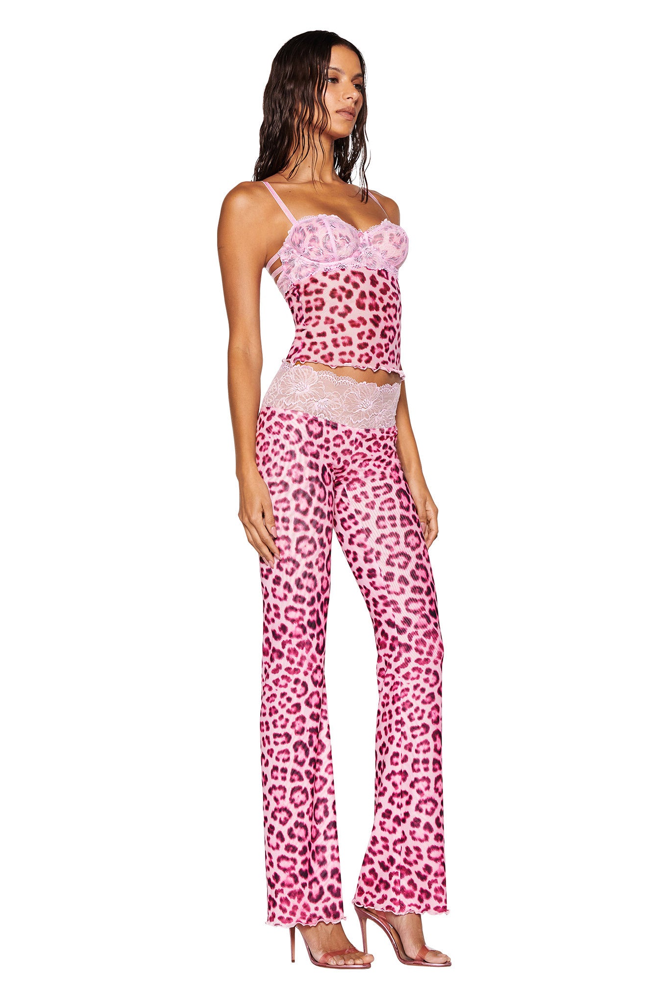 A woman showcases the SILO TOP in pink leopard print from I.AM.GIA, part of a stylish two-piece ensemble. The outfit, featuring a bodycon fit, includes a spaghetti strap top paired with flared pants, both enhanced with lace trim. She poses in profile against a plain white background while wearing high heels.
