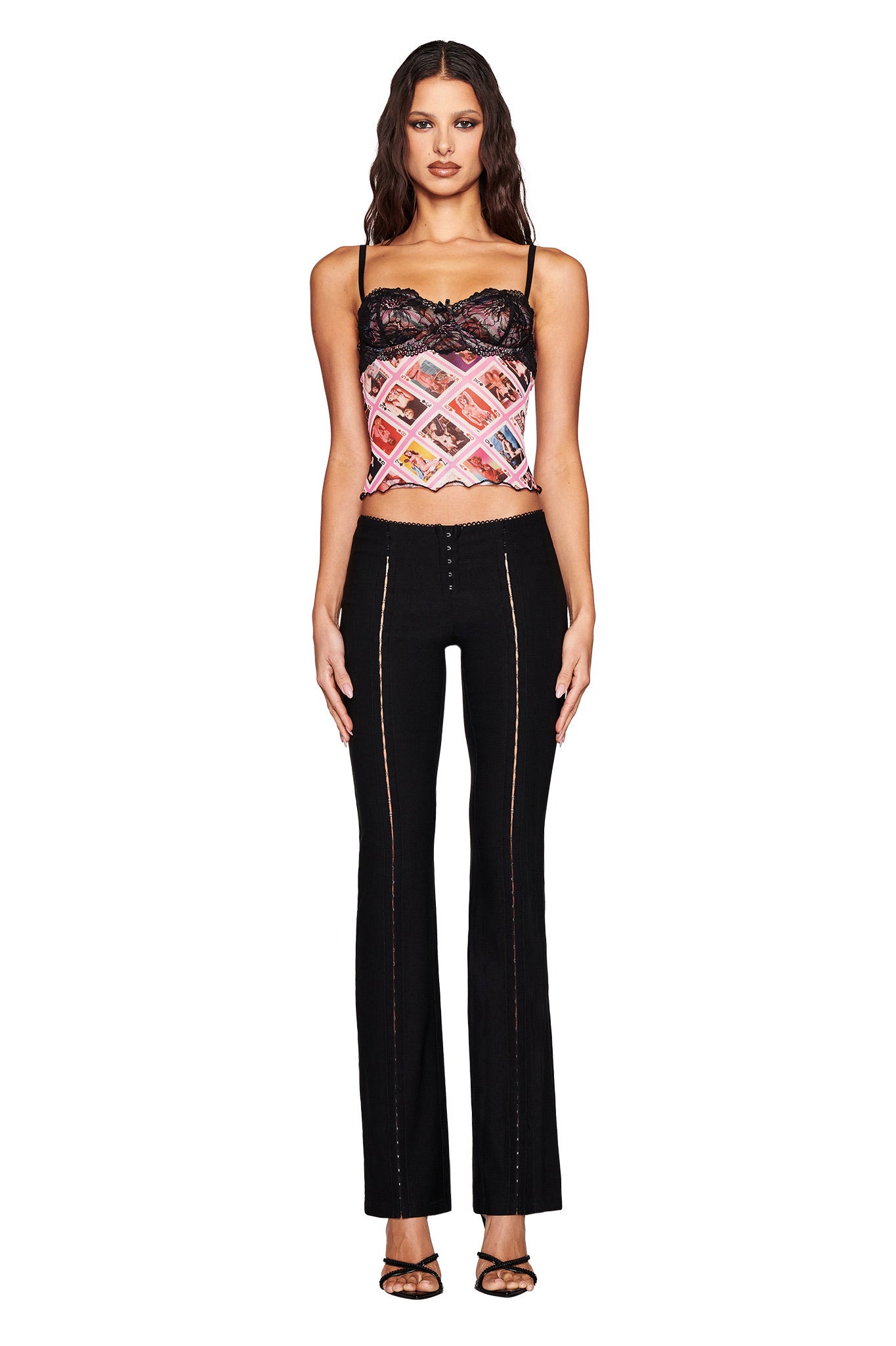 A woman with long dark hair confidently wears the SILO TOP - MULTI: GIA FACE CARD by I.AM.GIA, featuring black bustier lace and thin straps, paired with high-waisted black pants and open-toed heels. She poses forward against a white background.