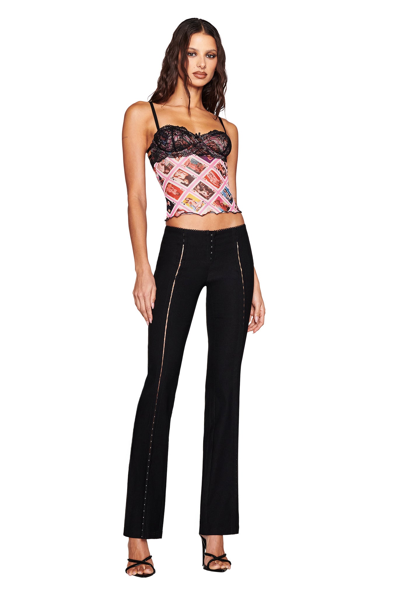 A woman confidently poses with her hand on her hip, wearing the I.AM.GIA SILO TOP - MULTI: GIA FACE CARD and black seam-design pants. Her chic ensemble is completed by heeled sandals and loose waves in her hair.