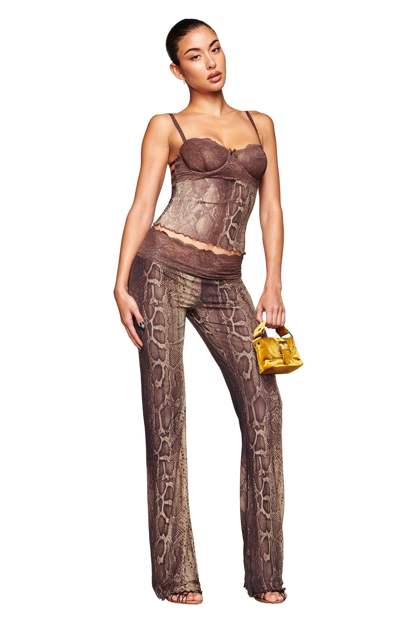 A woman models the I.AM.GIA SILO TOP - SNAKE, a brown snake print lace camisole with chic mesh bustier detail, paired with pants and a small yellow handbag, posing confidently against a white background.