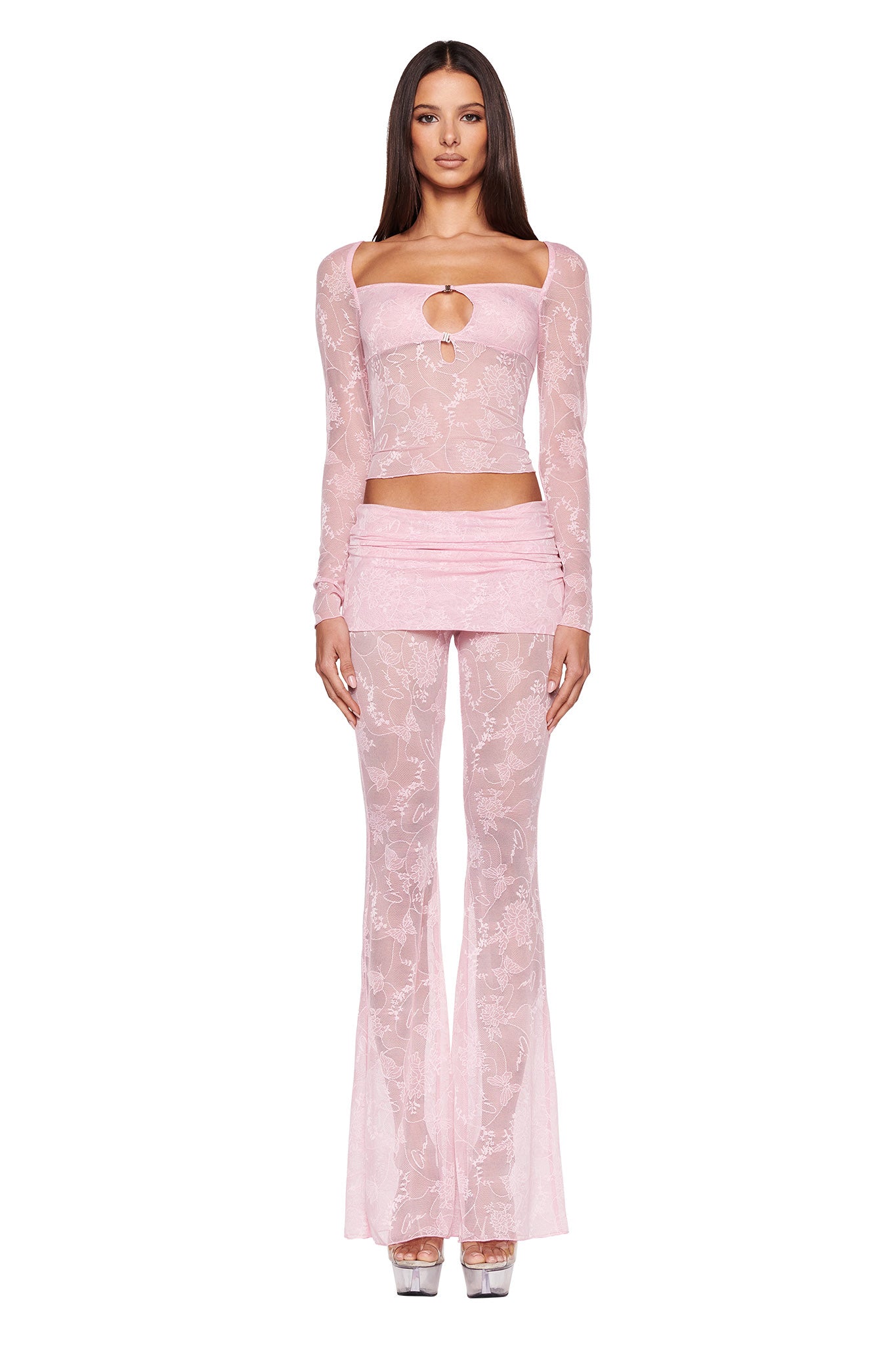 A woman stands facing forward wearing a pink, semi-sheer, long-sleeved I.AM.GIA top with floral patterns and a keyhole cutout. She pairs it with matching NALLIAH PANT - PINK : LACE from I.AM.GIA and silver platform heels.