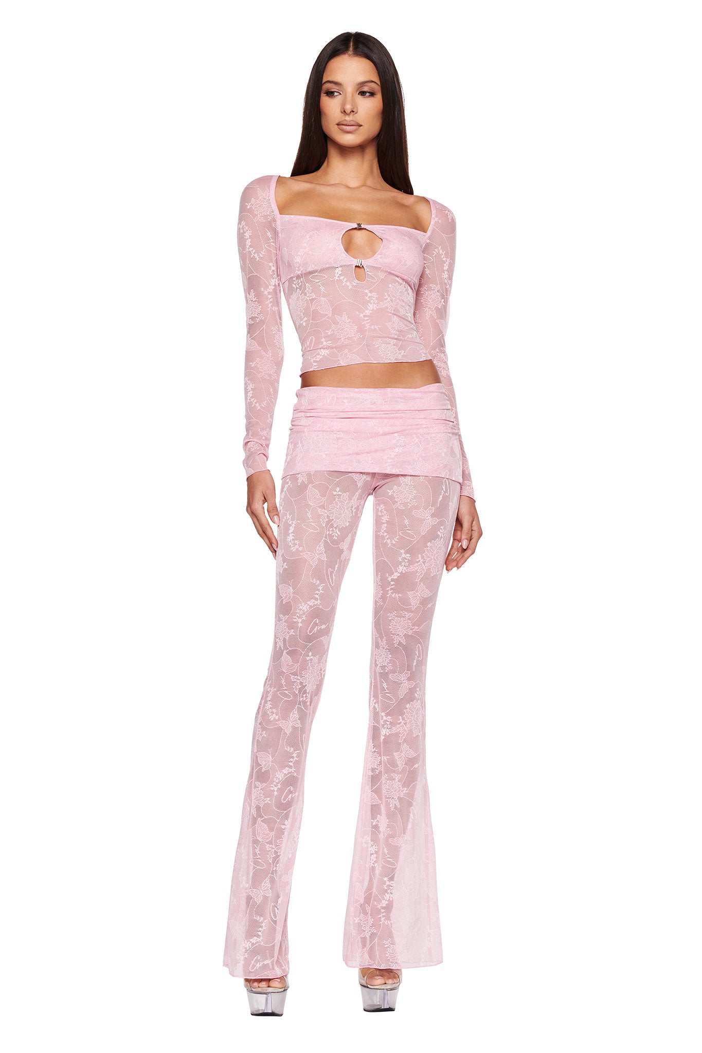 A woman models an I.AM.GIA NALLIAH PANT - PINK : LACE outfit. The ensemble includes a sheer pink lace long-sleeve crop top with a small keyhole cutout and matching high-waisted pants that flare into bell-bottoms. She stands facing forward, wearing high-heeled sandals.