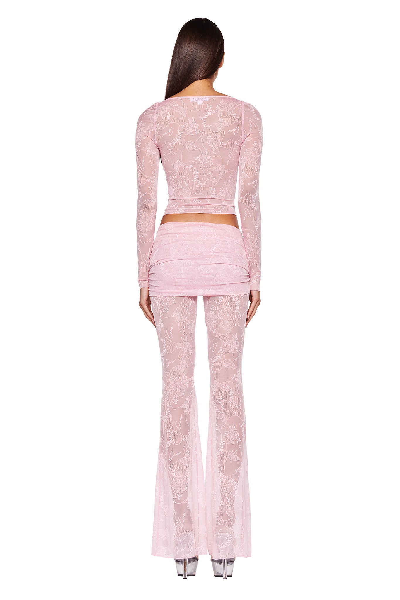 A woman wearing the NALLIAH PANT - PINK : LACE from I.AM.GIA, a sheer, pink, long-sleeved top and matching pants with a folded mini skirt overlay adorned with a delicate floral pattern, is viewed from the back. She completes the look with clear platform heels.