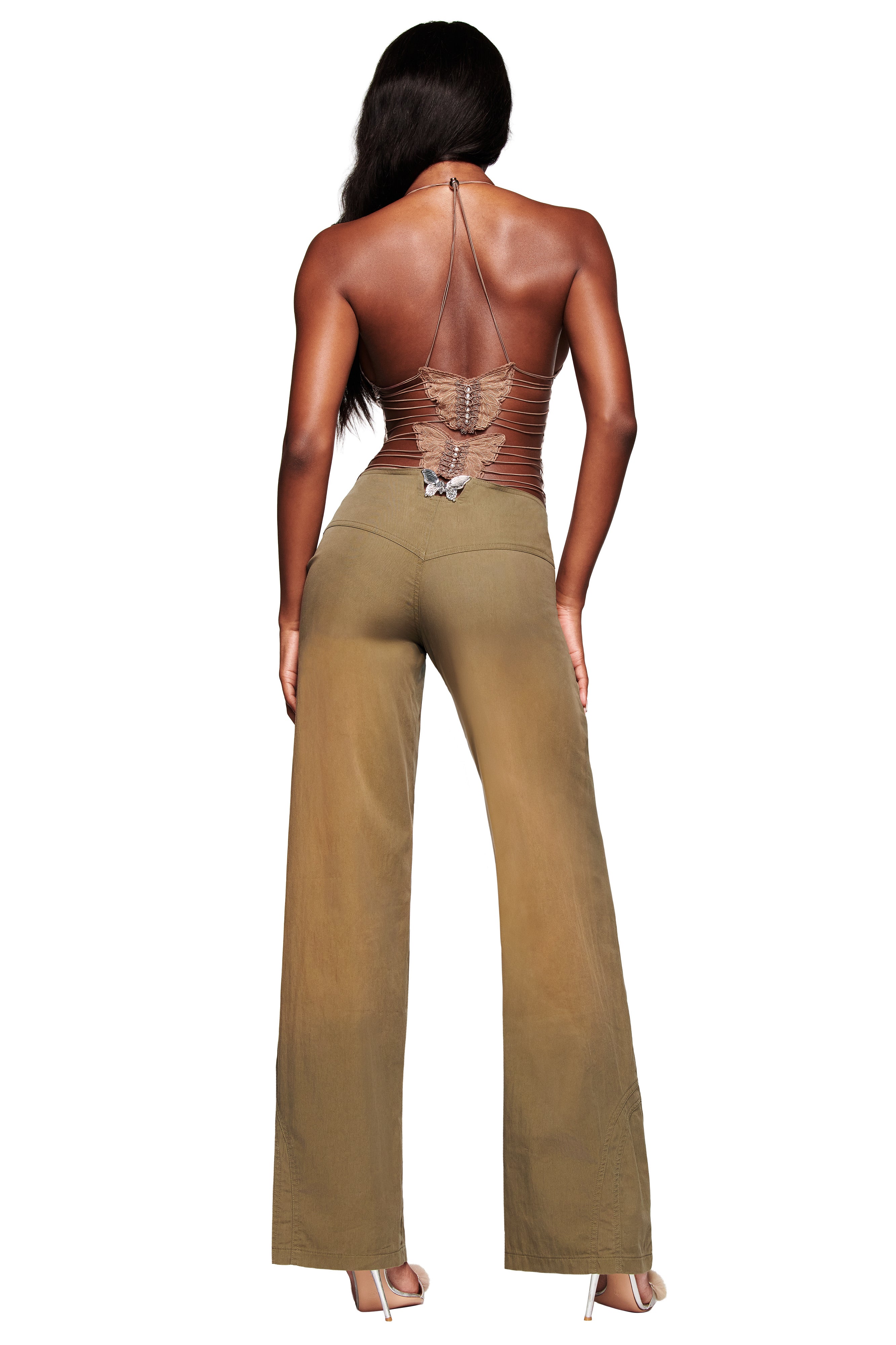 A woman with long dark hair faces away, wearing tan wide-leg pants and the I.AM.GIA ELLERY TOP - LEOPARD, an open-back strappy mesh halter. She completes the look with high heels against a white background.