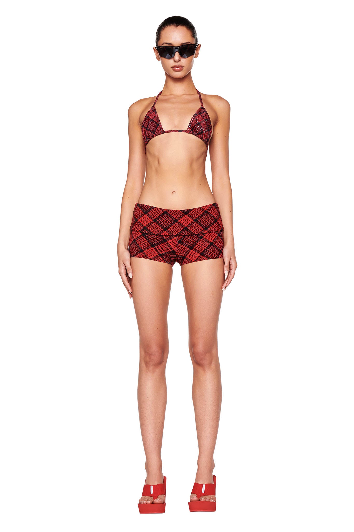 A woman stands facing forward, wearing I.AM.GIA sunglasses, the MARTINA BIKINI TOP - RED PLAID from I.AM.GIA, matching red plaid booty shorts, and red platform sandals. The outfit features a coordinated red plaid design. The background is plain white.