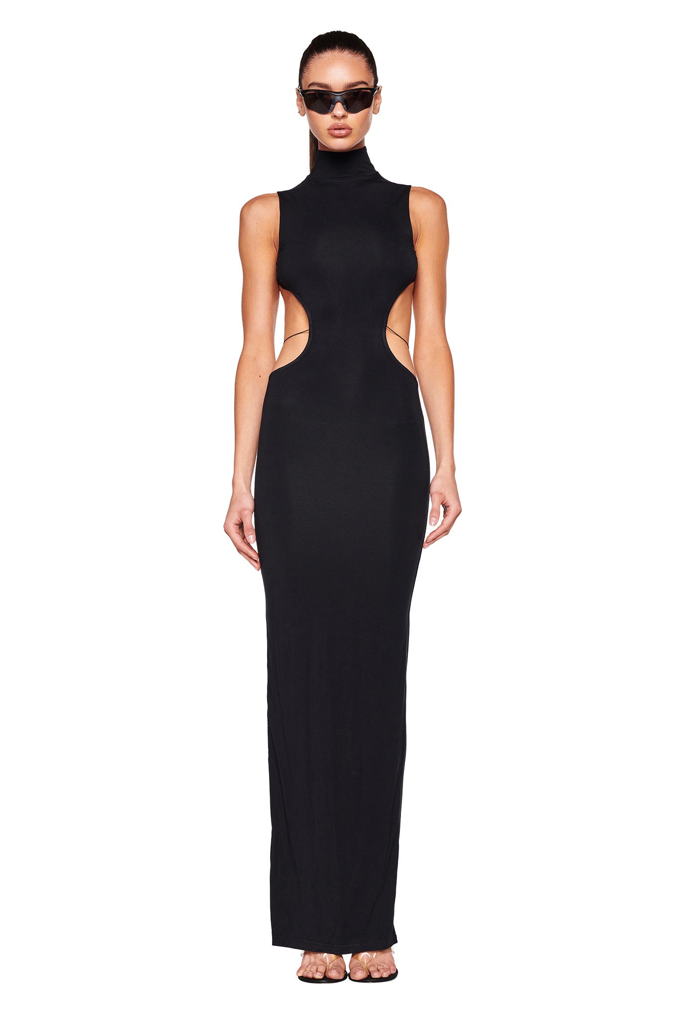 A woman stands in a form-fitting, sleeveless I.AM.GIA LARISA MAXI DRESS in black, featuring cut-out sides and a high neckline. Her hair is pulled back, she wears dark sunglasses, and her arms are relaxed at her sides. She completes the look with clear-strap high-heeled sandals against a white background.