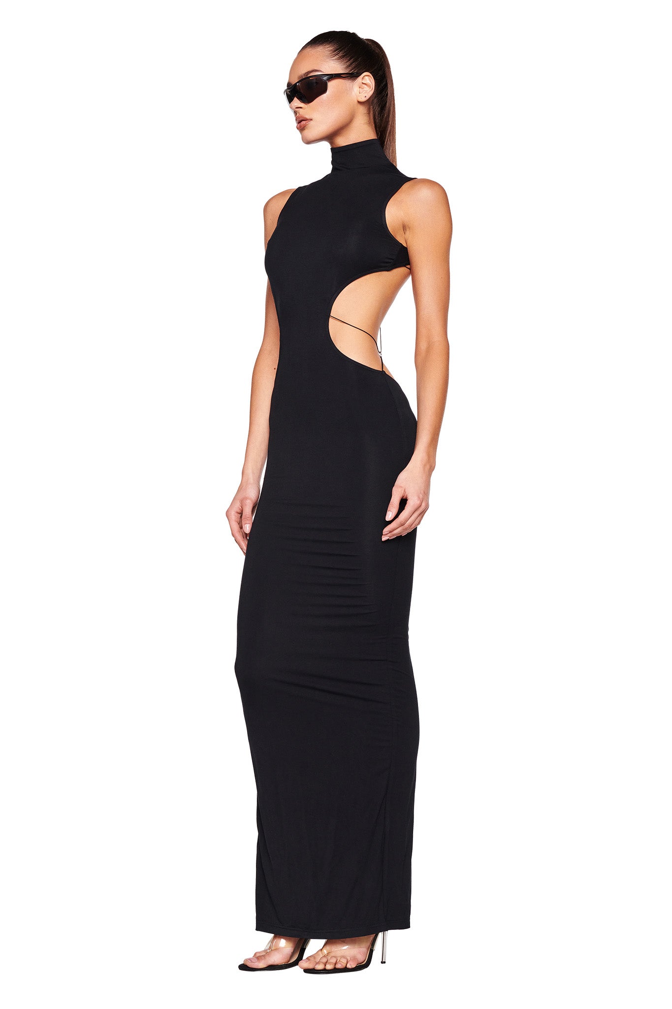 A woman stands confidently in an I.AM.GIA LARISA MAXI DRESS - BLACK, featuring a sleek, sleeveless design with side cut-outs and a high neckline. She accessorizes with black sunglasses and high-heeled sandals. Her hair is pulled back in a high ponytail, and she poses against a plain white background.