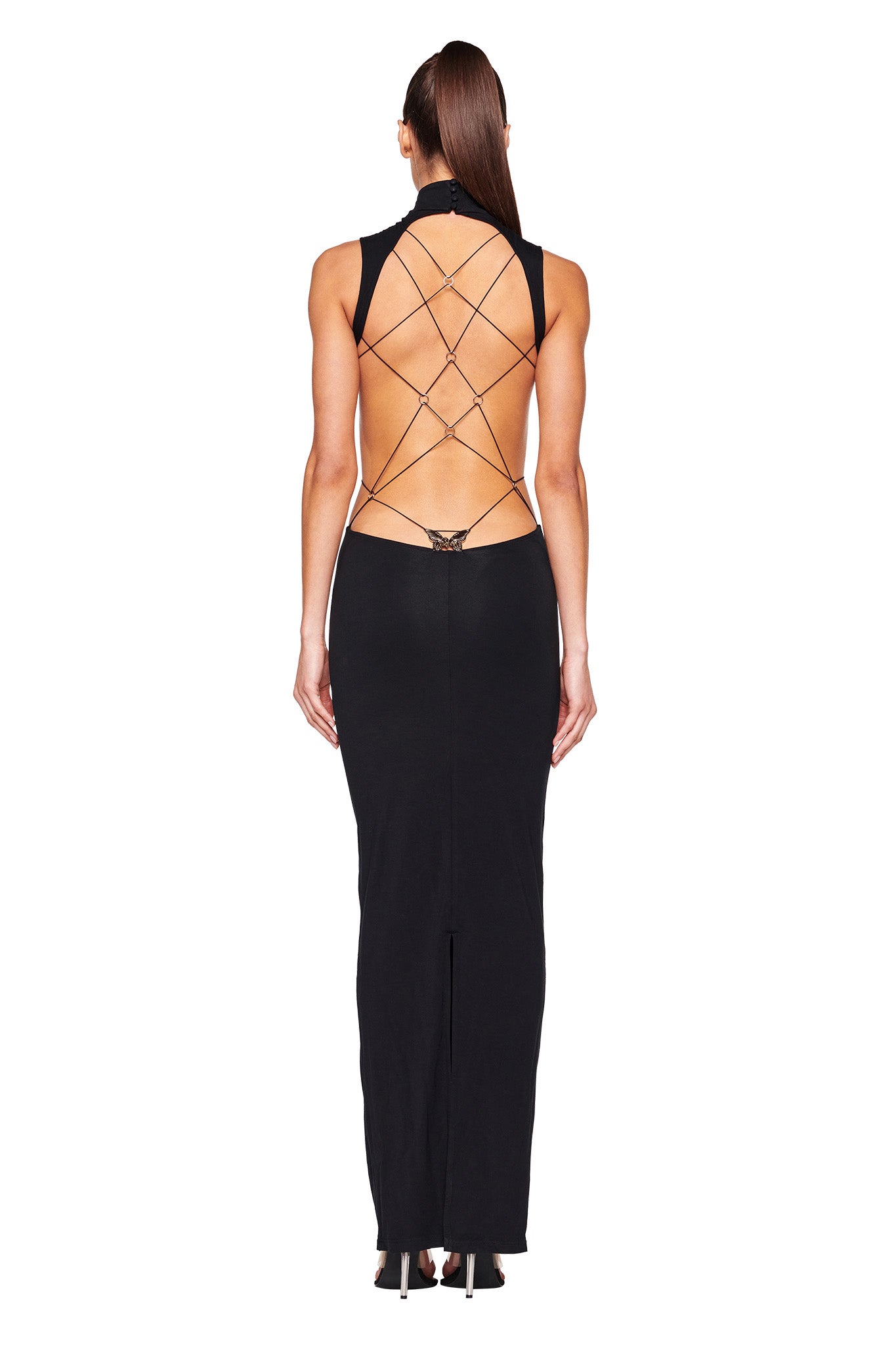 A woman is wearing the LARISA MAXI DRESS - BLACK by I.AM.GIA, featuring an open back and criss-cross strap design. The dress has a fitted silhouette and reaches the floor. She is facing away from the camera, showcasing the intricate back detail. Her hair is tied back.
