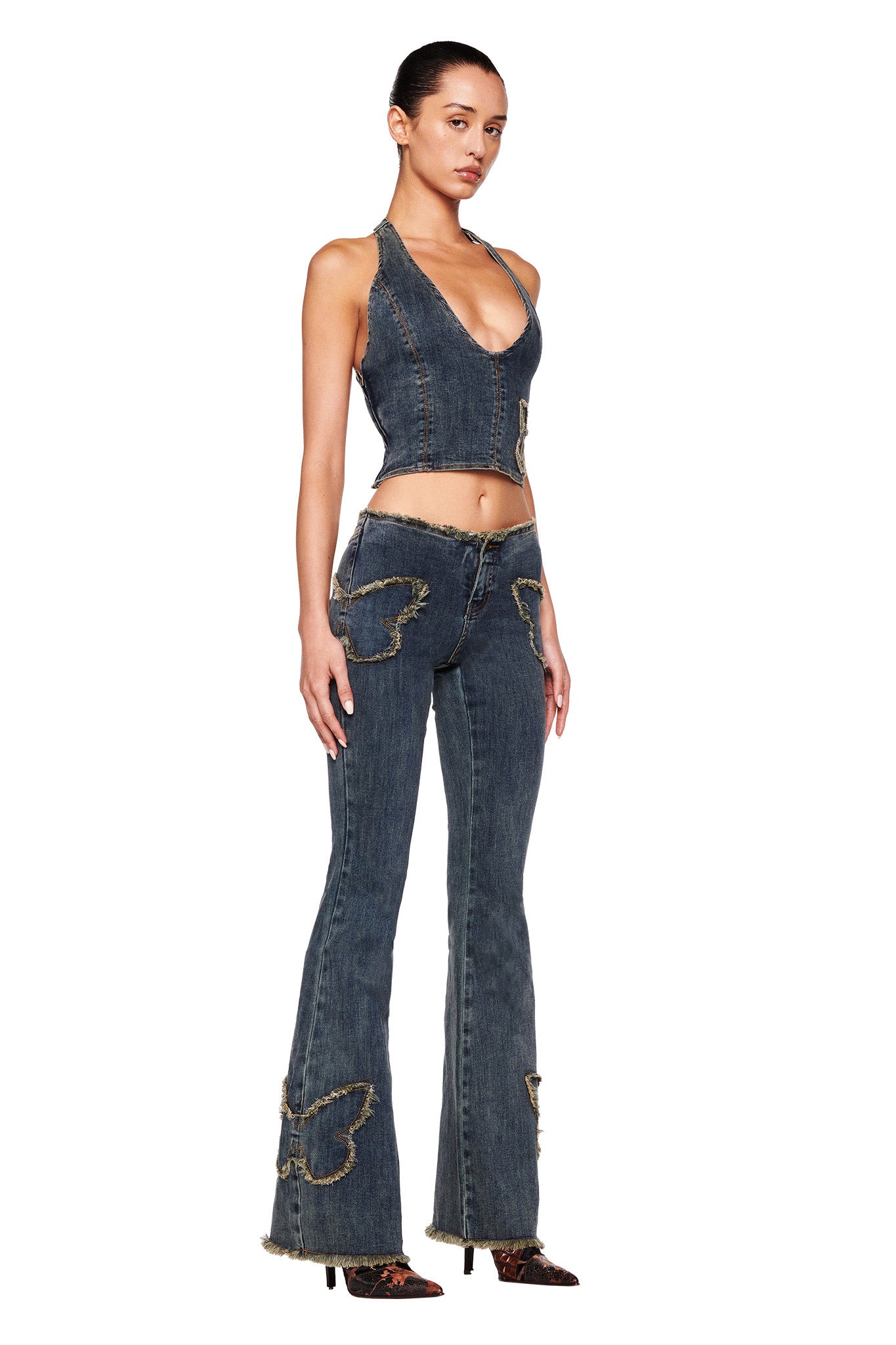 A woman stands facing slightly to her left, wearing a sleeveless stretch denim top with frayed edges. She pairs it with the I.AM.GIA SAFFIYAH PANTS in Denim Blue, high-waisted flare jeans featuring distressed butterfly patches. Her hair is pulled back, and she completes the look with black pointed-toe heels.