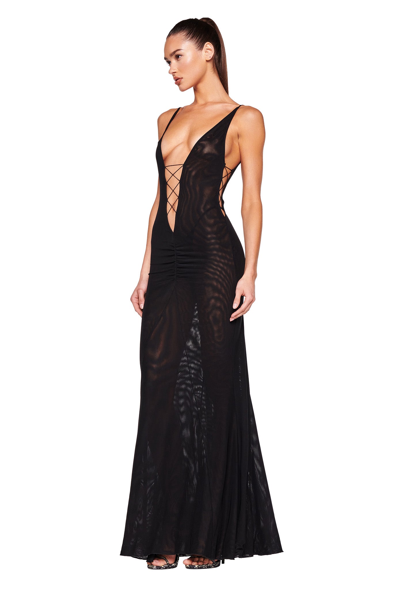 A woman stands in a fitted MAYA DRESS - BLACK by I.AM.GIA, featuring a deep plunging neckline with crisscross lacing, side cutouts, and a sheer flowing skirt. She poses with her right hand on her hip and her hair styled in a sleek high ponytail.