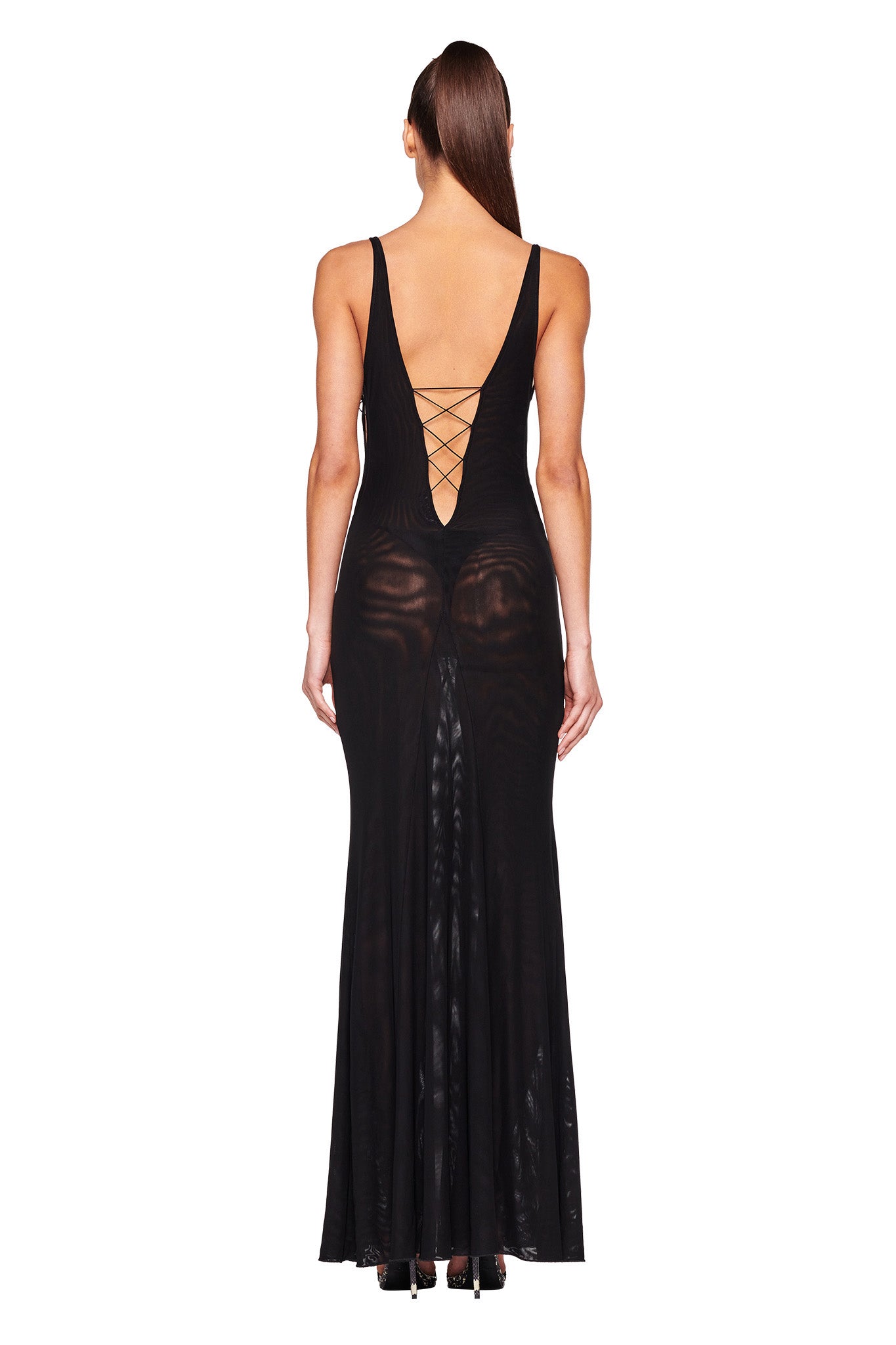 A woman is standing with her back to the camera, showcasing the MAYA DRESS - BLACK by I.AM.GIA. The floor-length dress features a deep V-shaped back with crisscross string detailing. She has long, straight hair tied back in a sleek ponytail and is wearing high-heeled shoes.