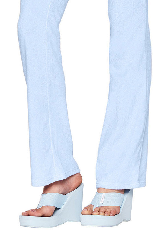 A person is wearing light blue I.AM.GIA pants that flare at the bottom, paired with the SHELBY FLIP FLOP - BLUE : BABY BLUE open-toe platform sandals. The sandals perfectly match the light blue pants. The photo focuses on the lower legs and feet, with a white, uncluttered background.