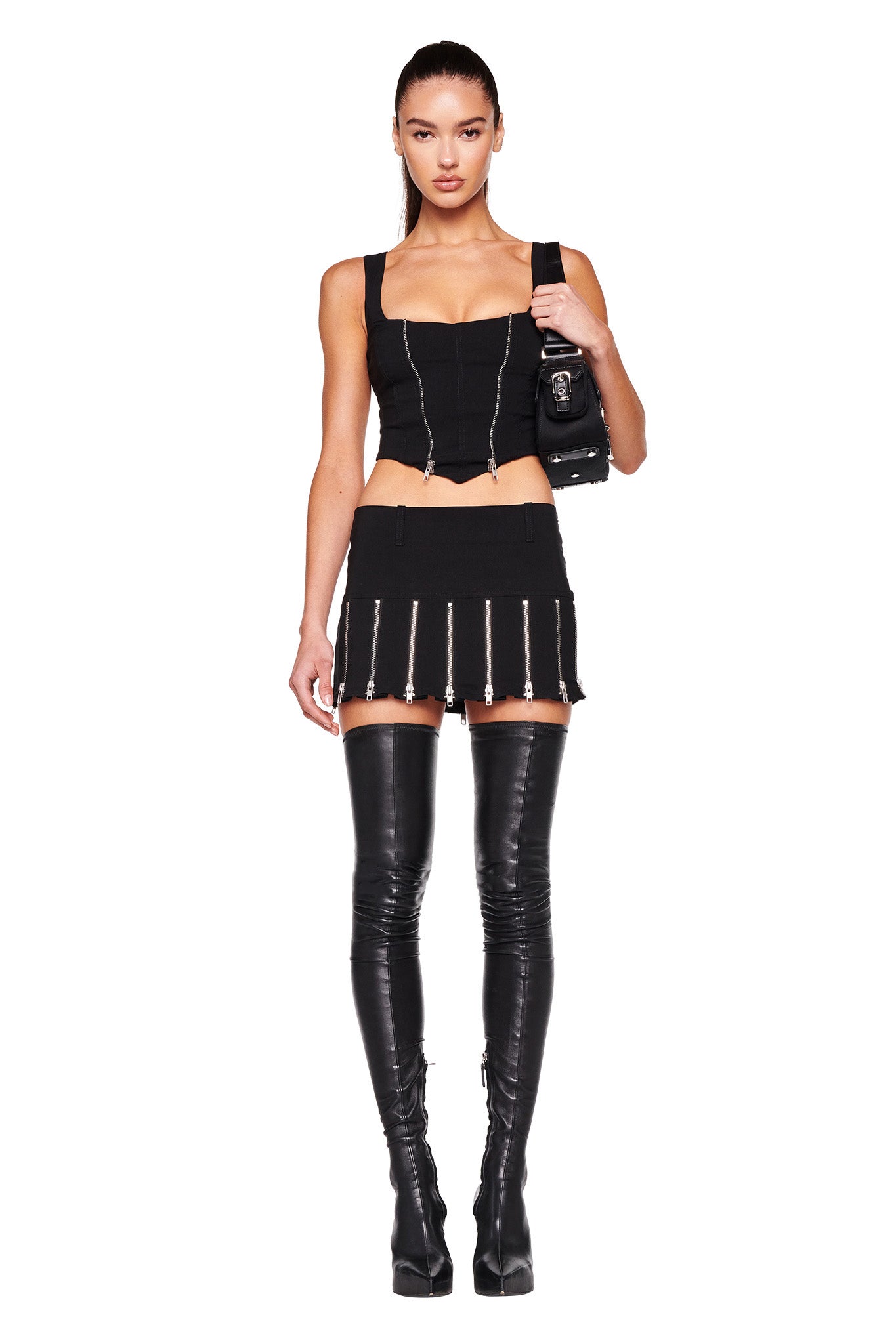 A woman stands with her hips slightly turned, showcasing her edgy I.AM.GIA black crop top paired with the stylish CLAUDIA SKIRT - BLACK from I.AM.GIA, accentuated by zipper details. Her outfit is completed with thigh-high black leather boots and a chic black shoulder bag. She has her hair tied back in a sleek ponytail against a plain white background.