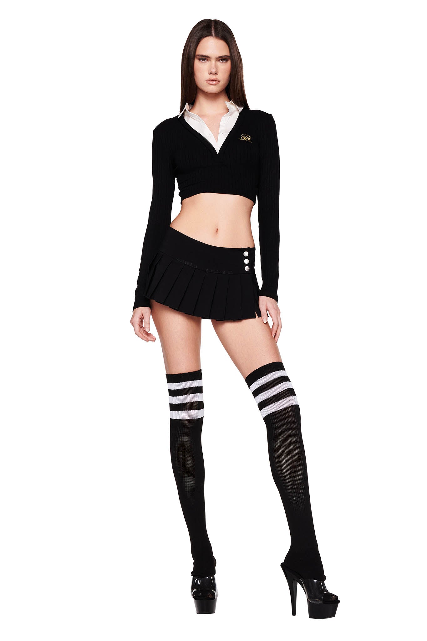 A person stands confidently wearing the LANDON TOP - BLACK from I.AM.GIA, along with a black mini skirt, and black thigh-high socks with white stripes. They are also wearing black high-heeled shoes. The background is plain white.