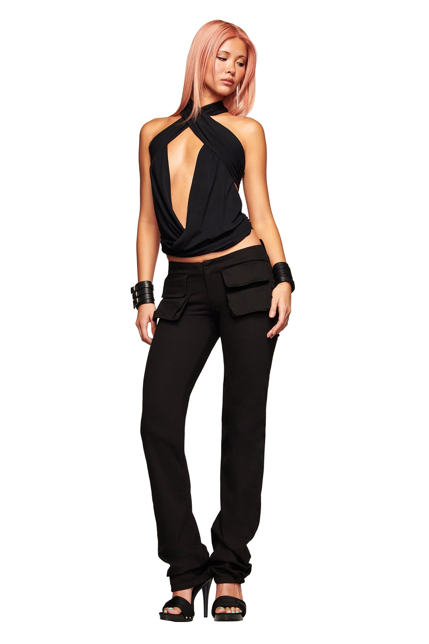 A woman with pink hair wears the I.AM.GIA KIMANI PANT in black, a sleeveless black halter top with a deep front cutout, black high heels, and black bracelets, standing against a white background.